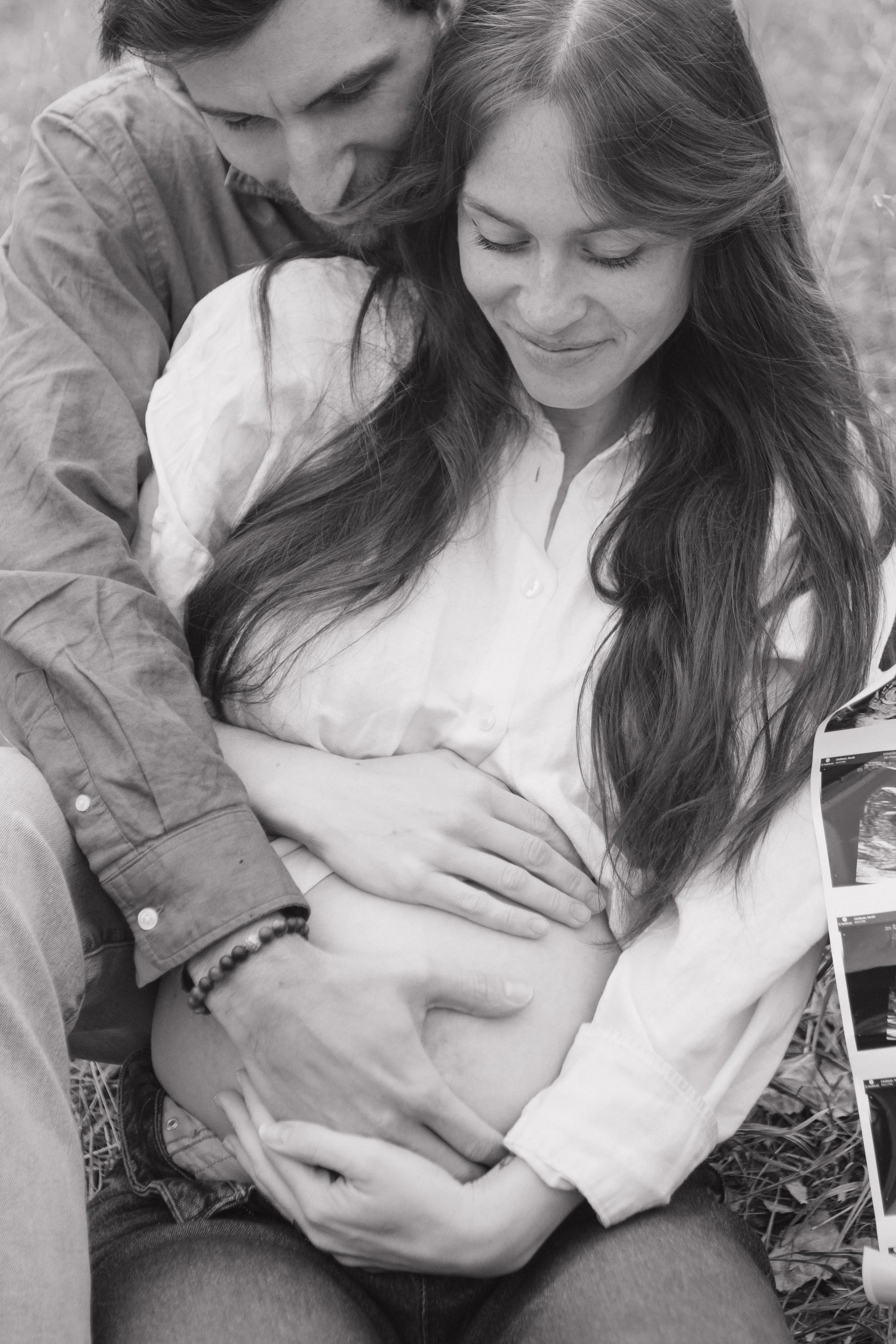 A pregnant woman with long hair sitting outside, smiling with her eyes closed as a man and her hand are on her belly, with ultrasound images nearby.