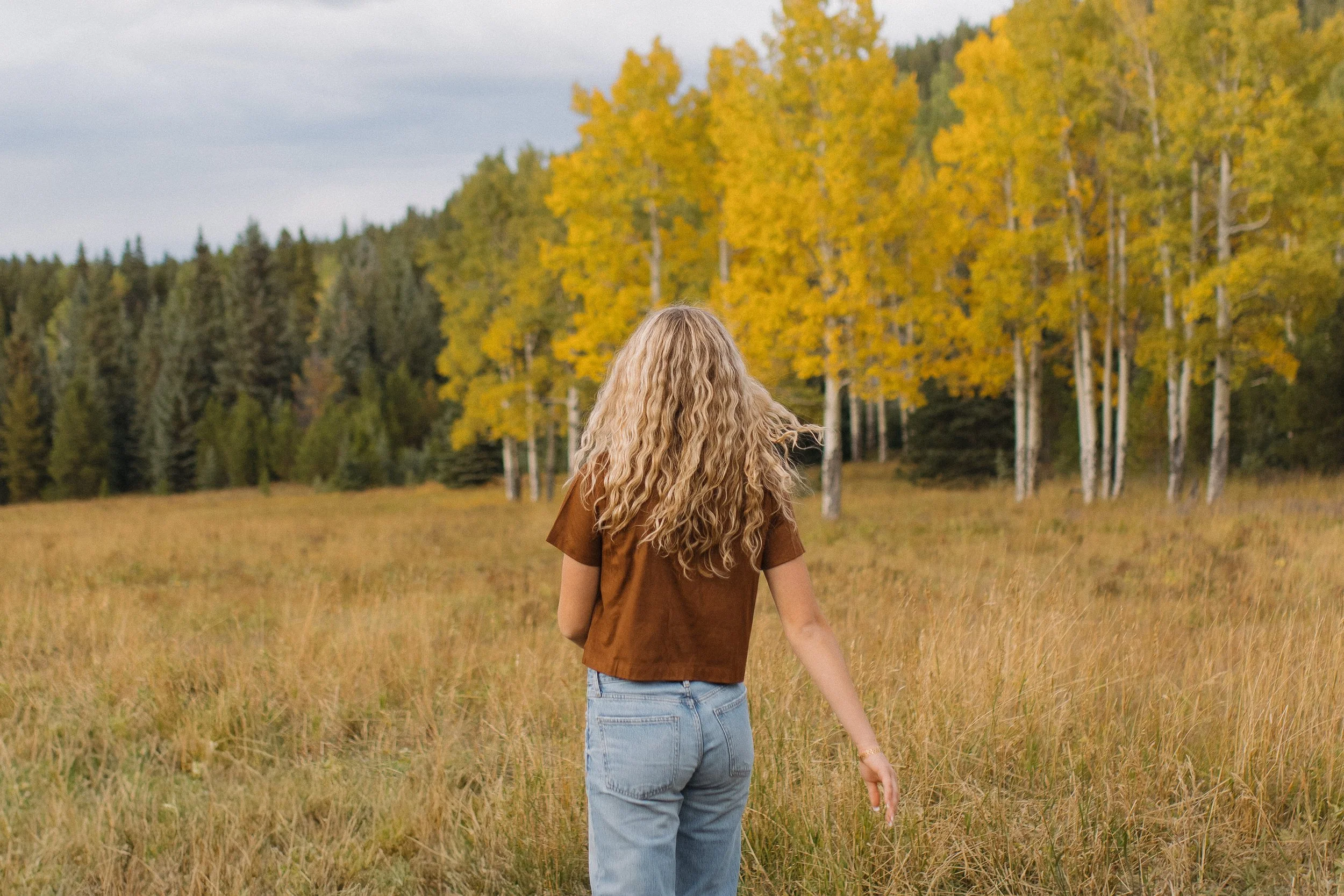 A woman with curly blonde hair walking through a grassy field in autumn, with a background of yellow and green trees under a cloudy sky.