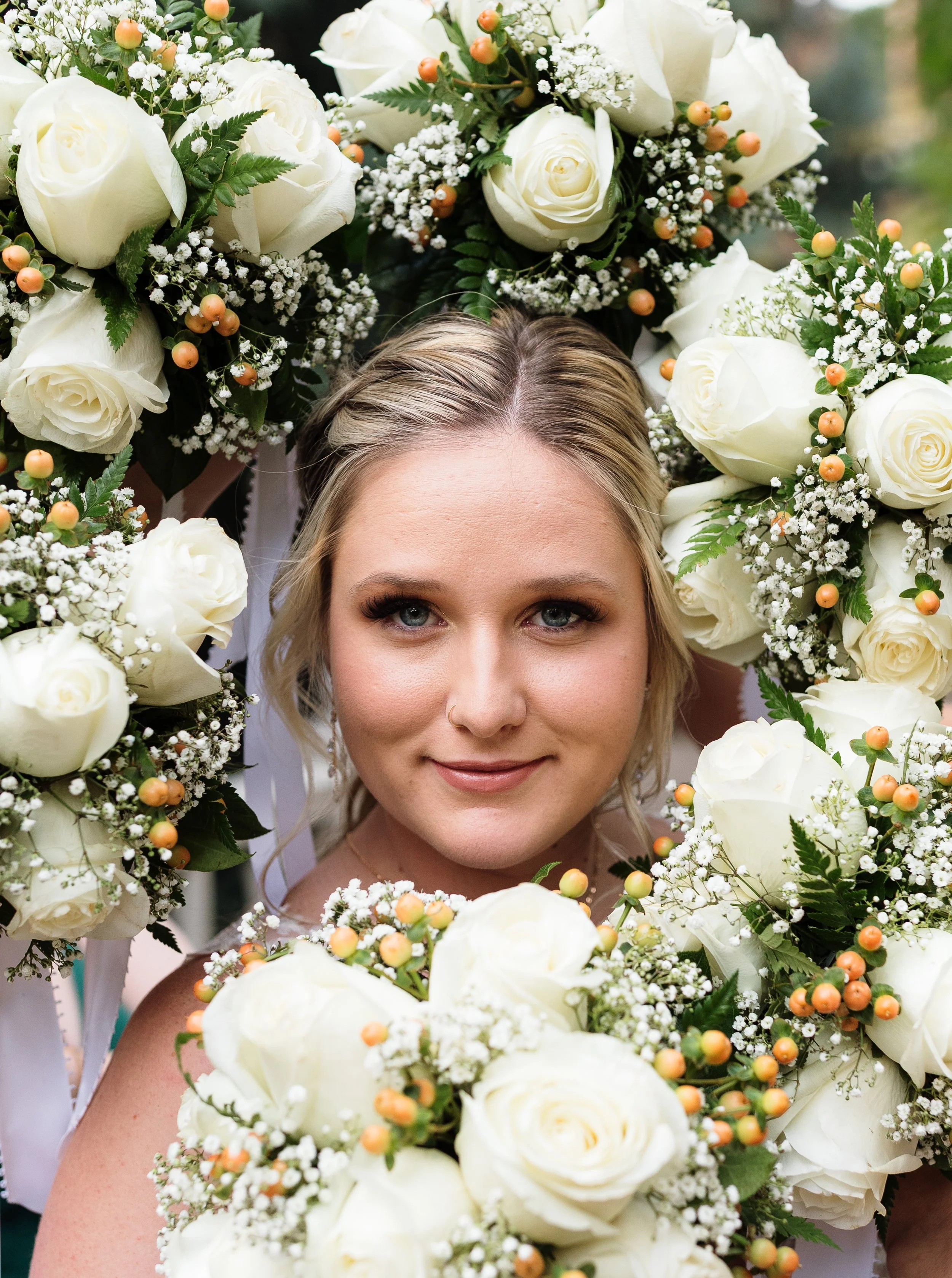 A smiling woman surrounded by large white roses and flowers, with greenery and small orange berries, forming a floral wreath.