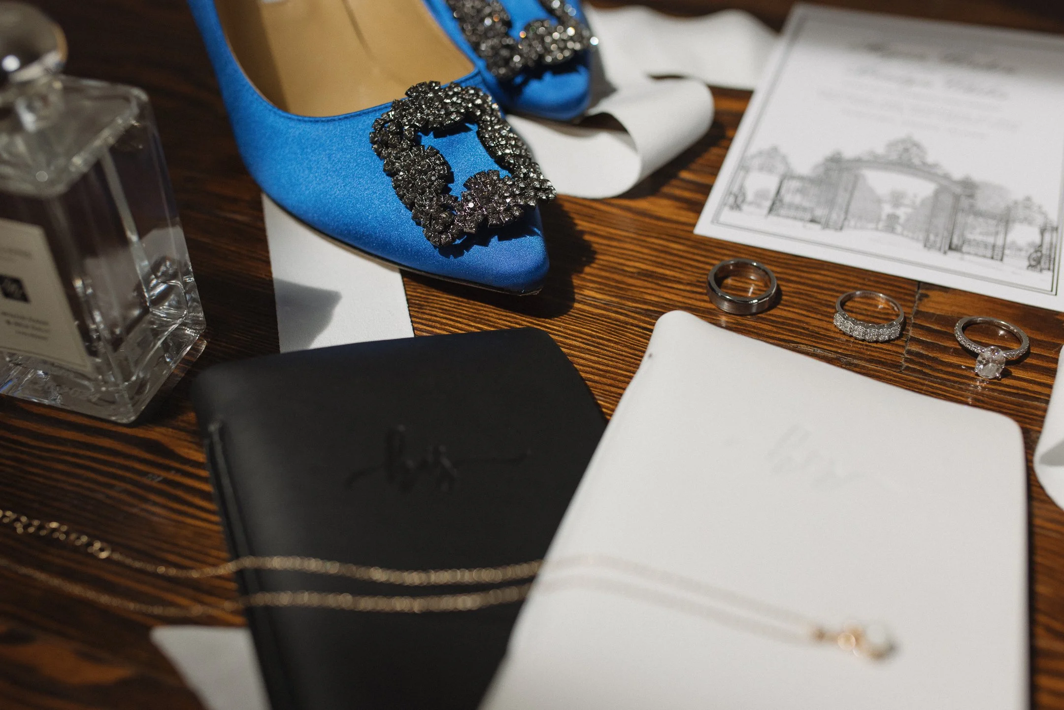 Blue satin shoes with large black rhinestone buckles, various jewelry including rings and a necklace, perfume bottle, and wedding invitation on a wooden surface.
