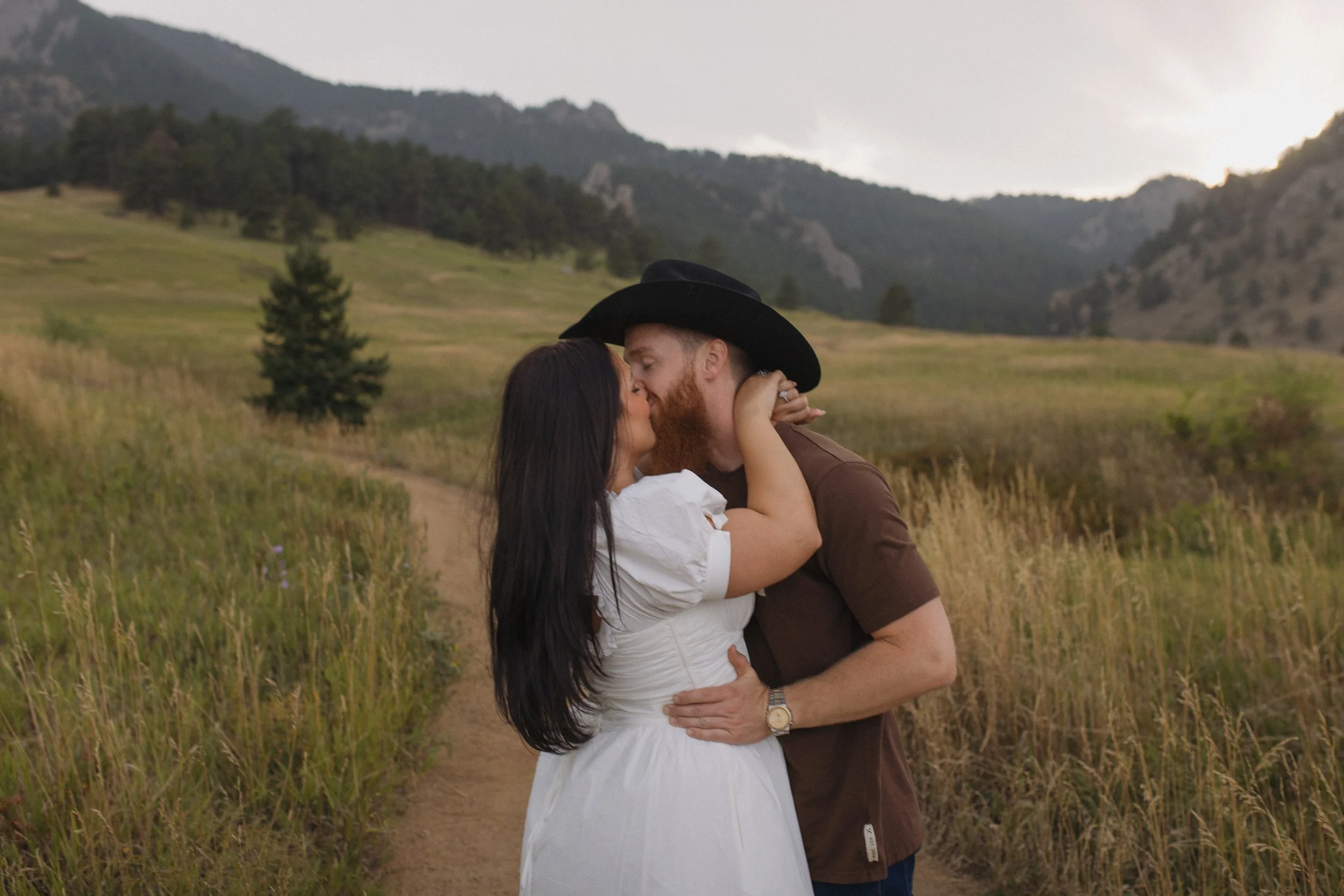 A couple kissing on a dirt path in a grassy outdoor setting with hills and mountains in the background during the rain.
