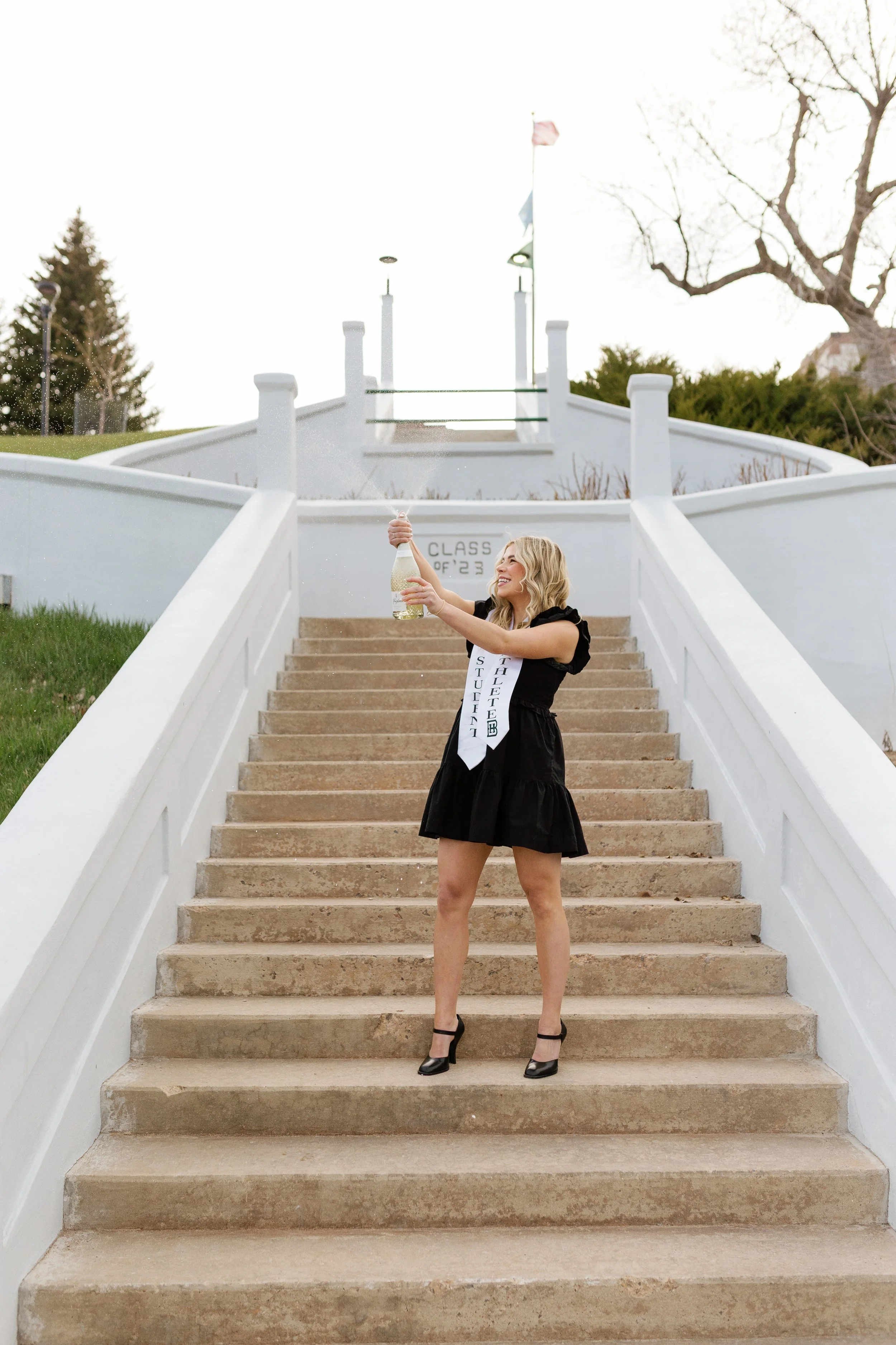 Young woman in a black dress and high heels celebrating on front steps of a school, holding a bottle of champagne.