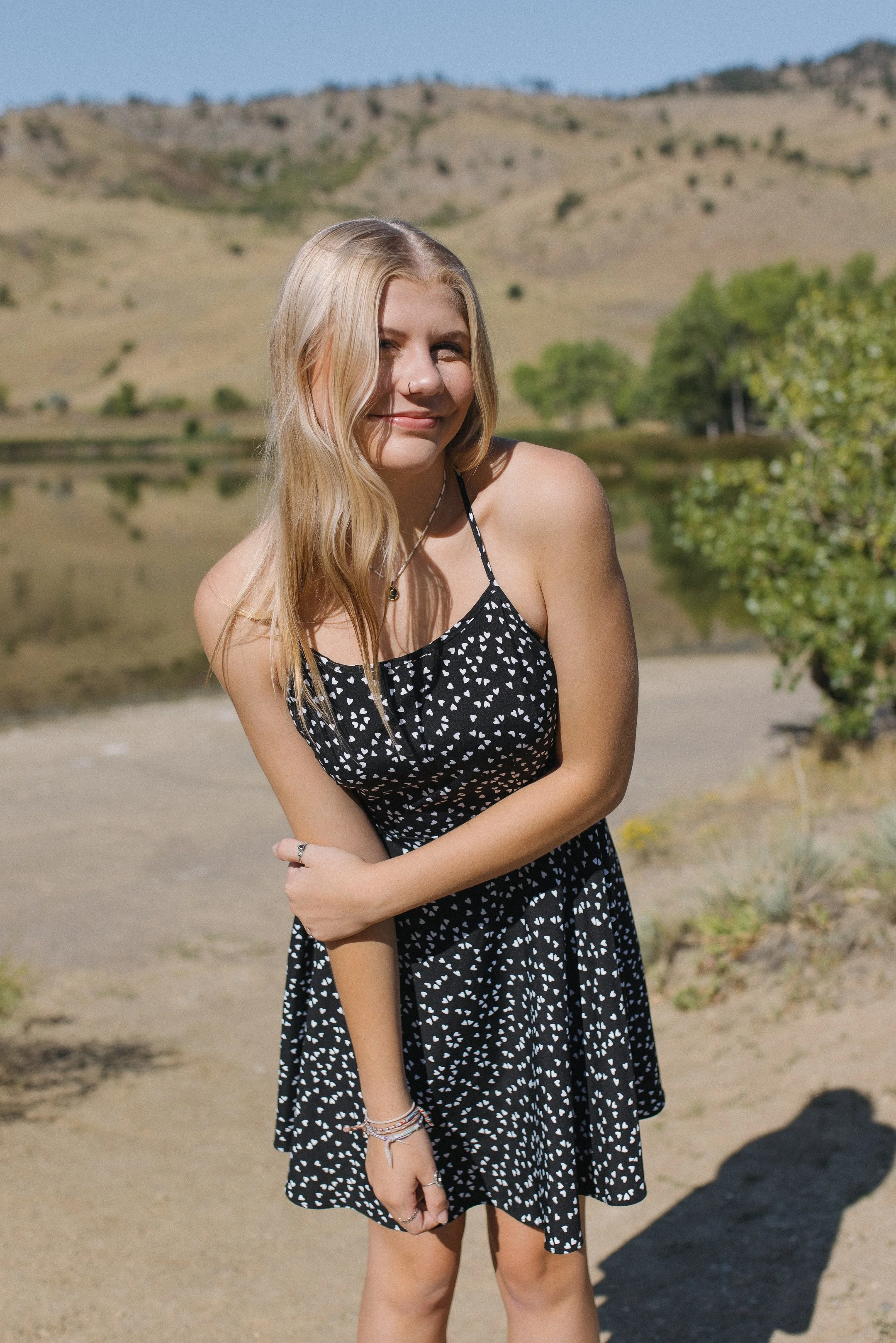 High school senior with long blonde hair wearing a black dress with white heart patterns stands outdoors near a lake, with hills and a partly cloudy sky in the background.