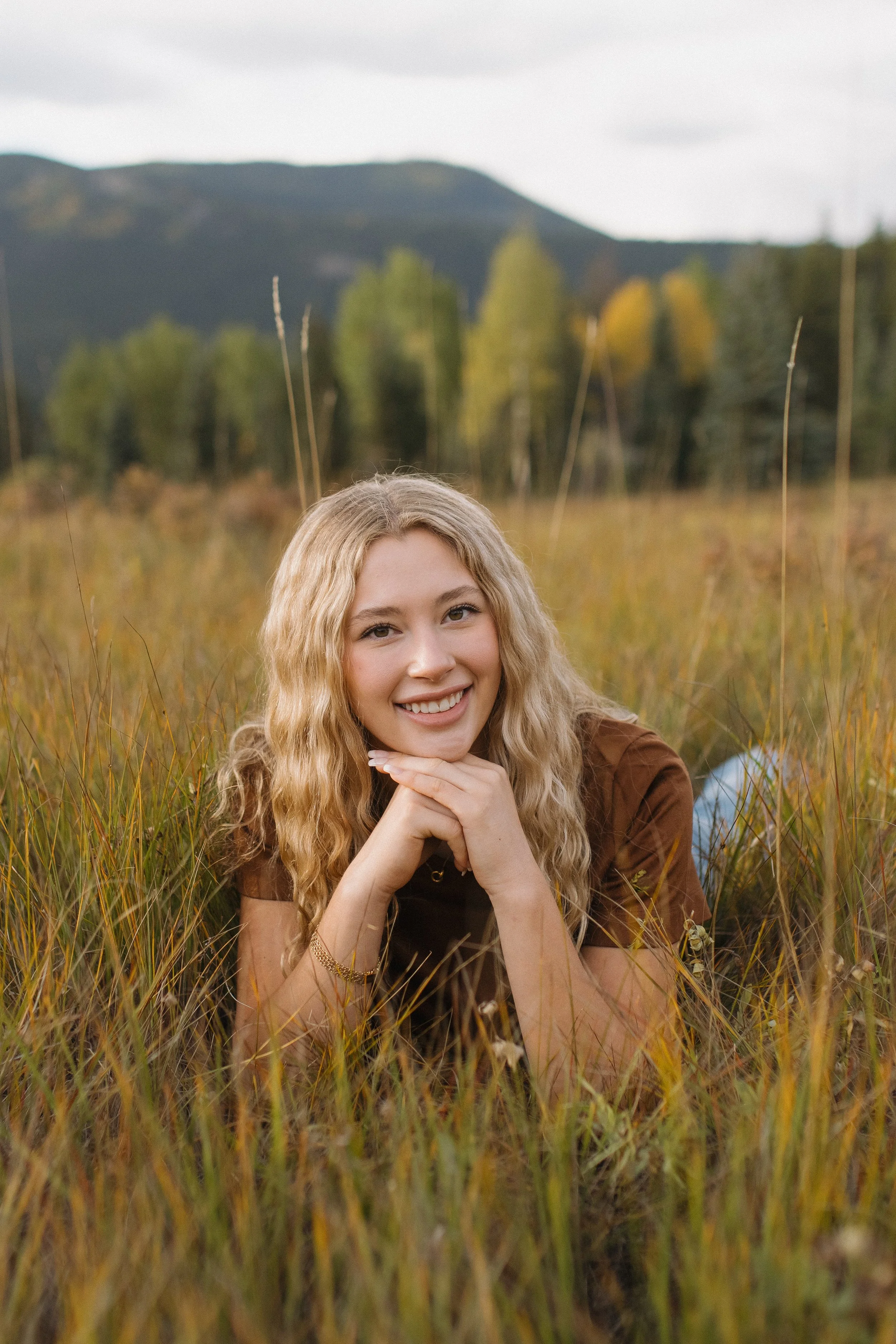 A young woman with curly blonde hair smiling and lying on her stomach in a grassy field, with mountains and trees in the background.