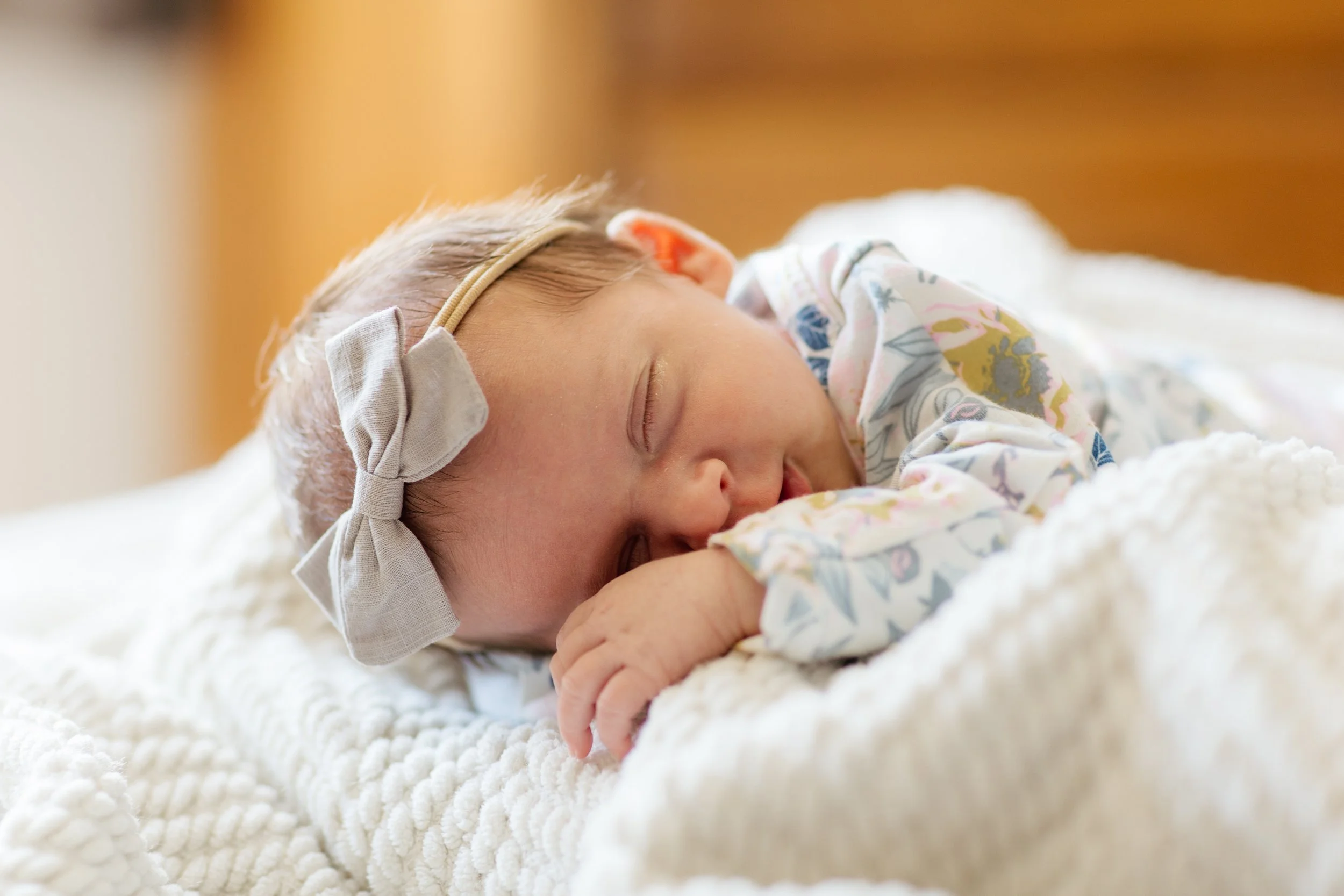 A sleeping baby girl lying on a soft, cream-colored blanket, wearing a headband with a bow and a floral outfit.