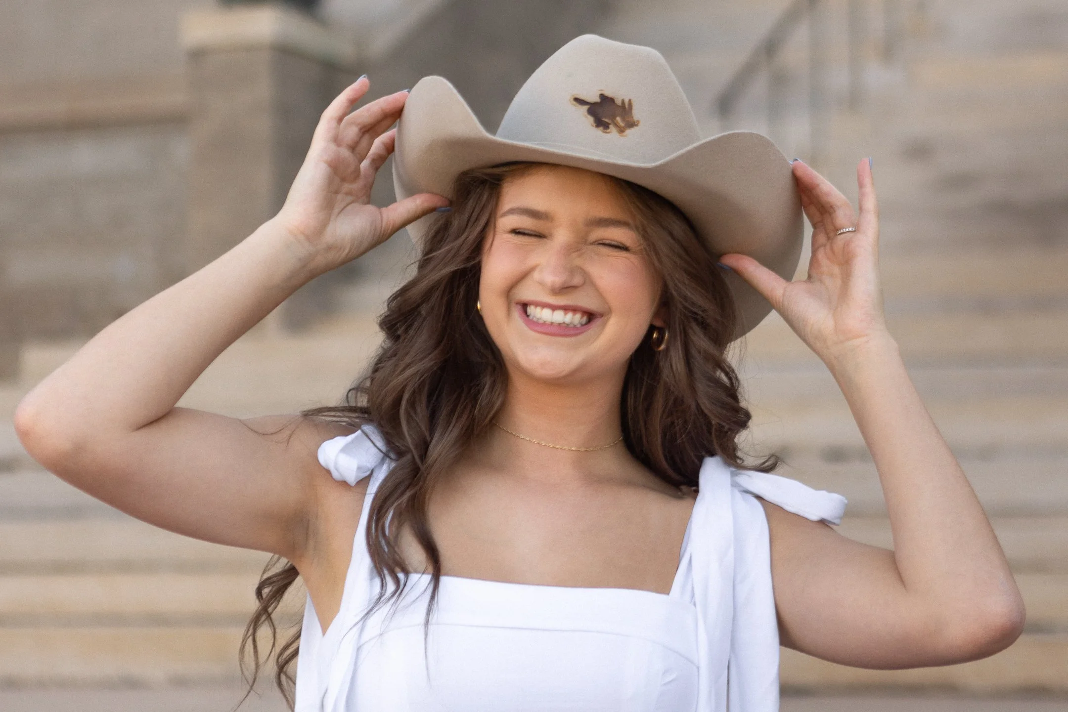 Graduation photos of a girl wearing a white dress with tied shoulders and a beige cowboy hat with a small animal design, outdoors near stone steps.