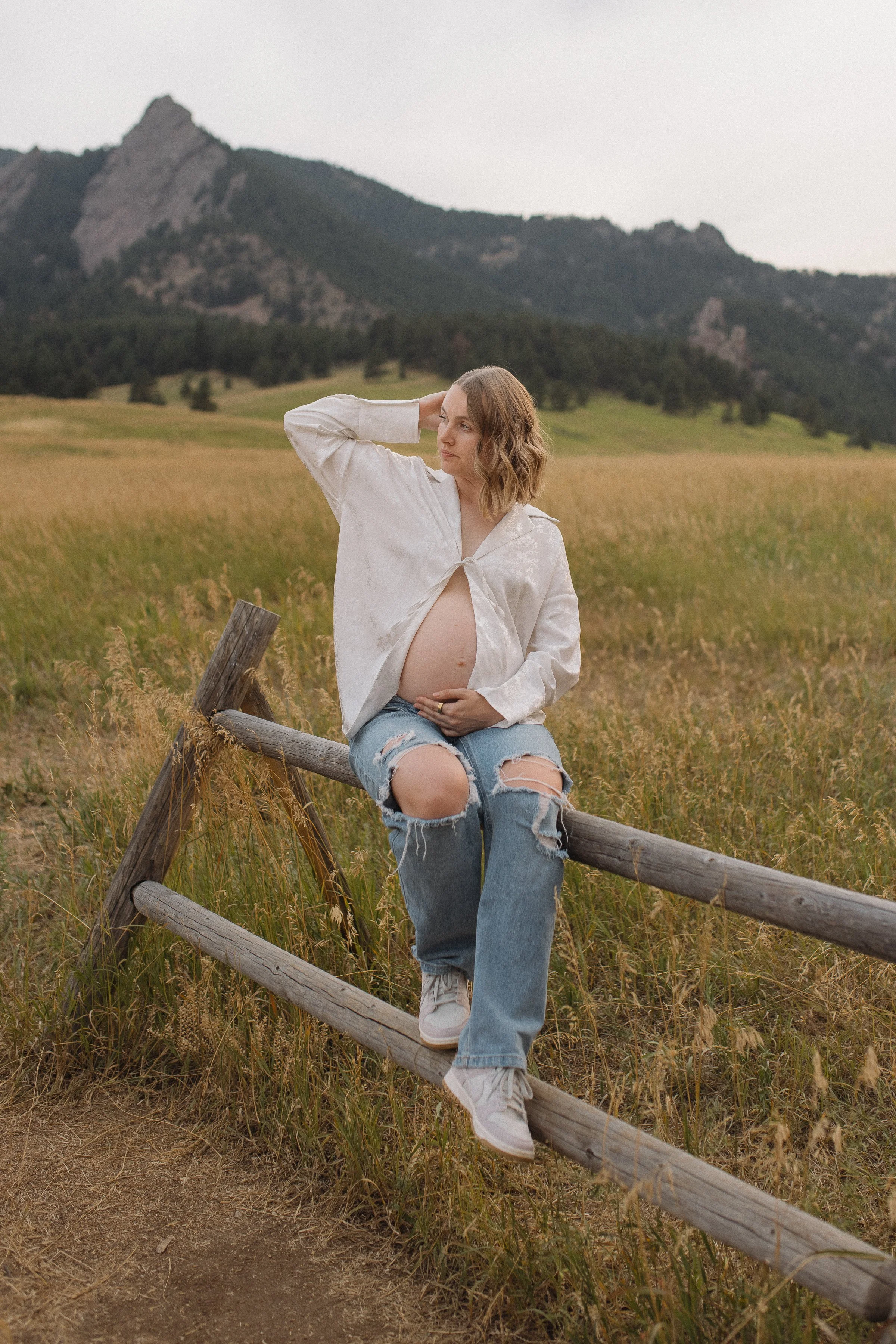 Pregnant woman sitting on a wooden fence in a grassy field with mountains in the background.