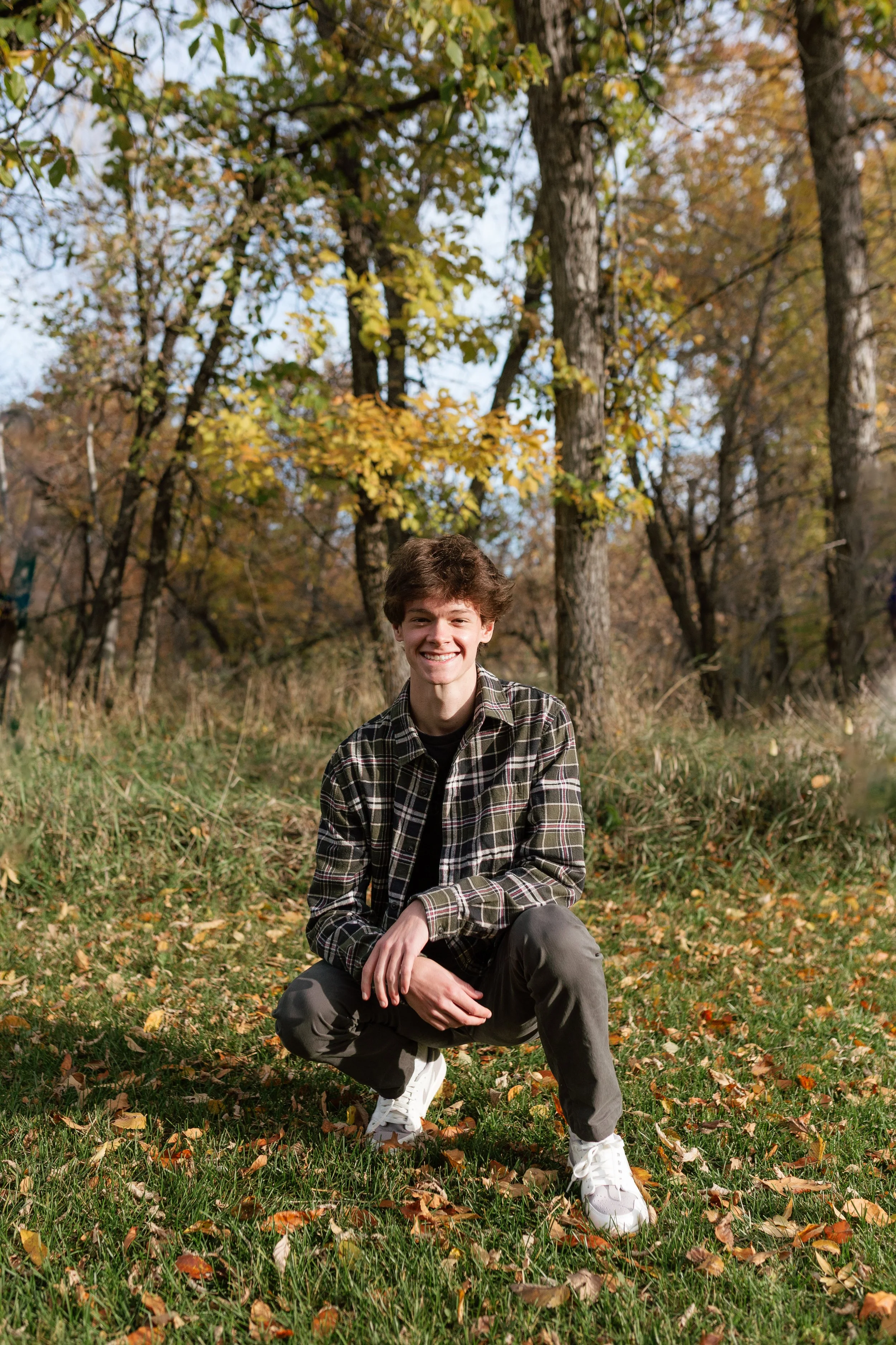 A young man with brown hair smiling and squatting outdoors on a grassy area covered with fallen autumn leaves, surrounded by trees with orange and yellow foliage.