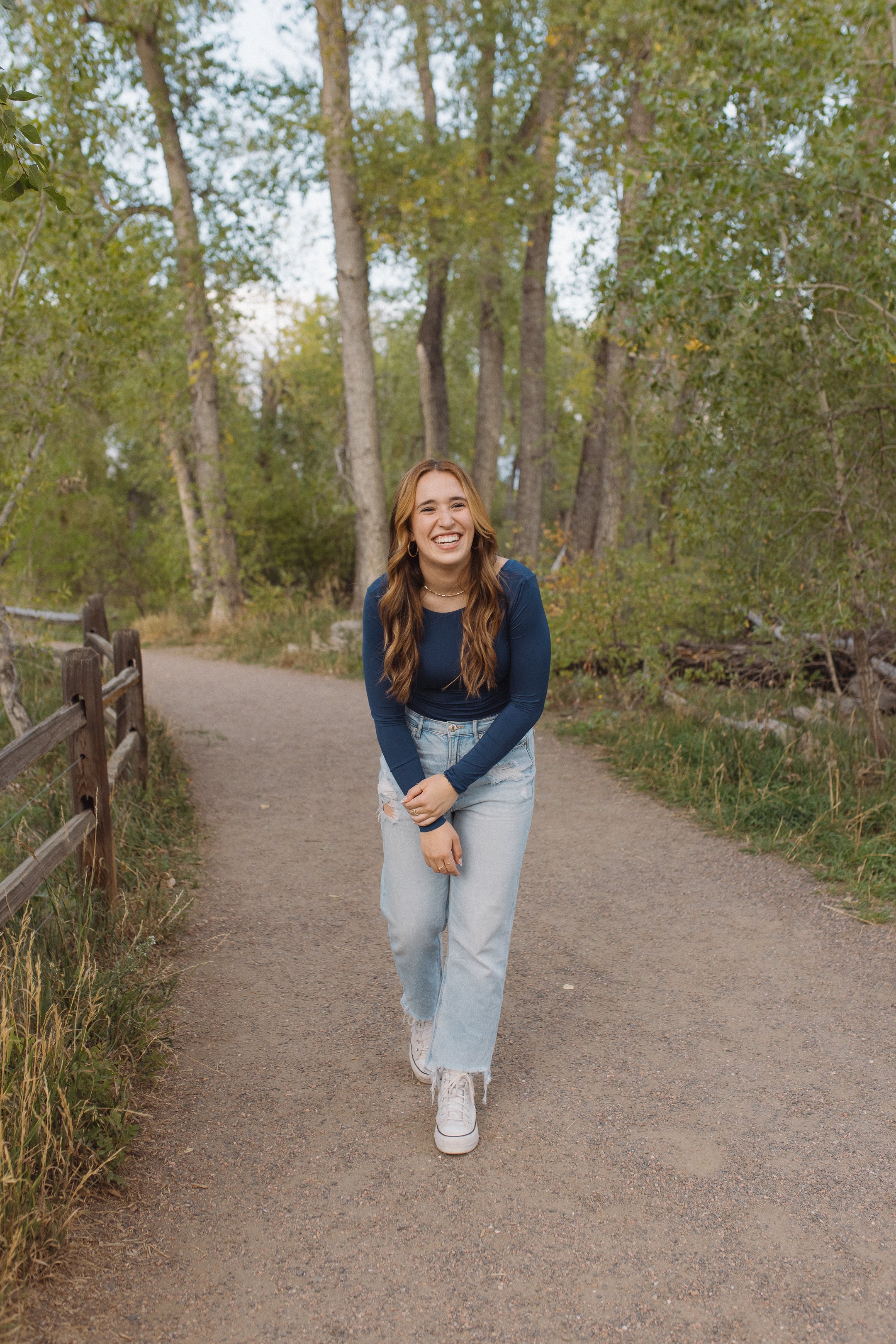 High school senior with long brown hair wearing a blue long-sleeve top, light blue jeans, and white sneakers standing on a dirt path in a wooded area, smiling and laughing.