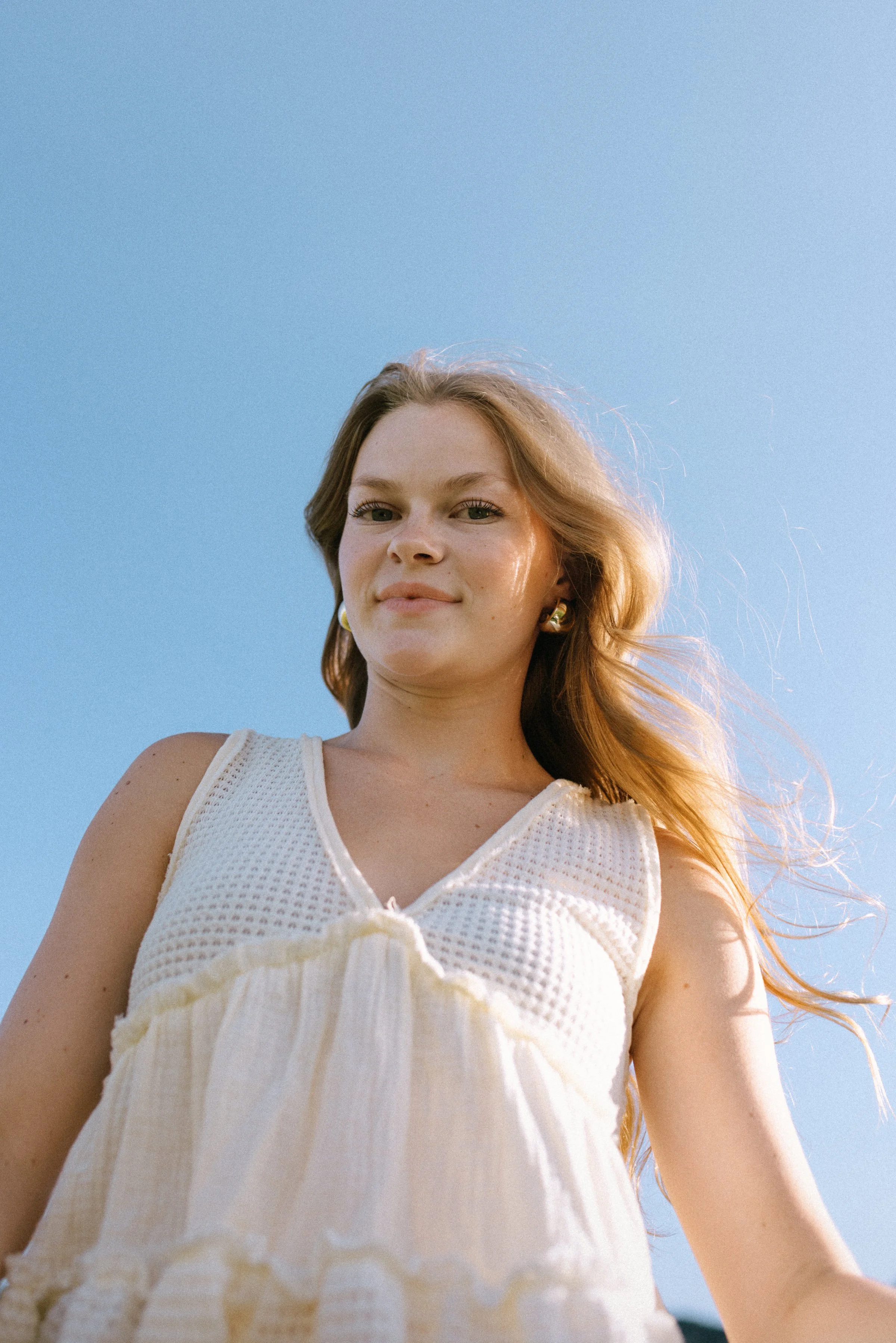 Young woman with long hair wearing a sleeveless white dress, standing outdoors against a clear blue sky.