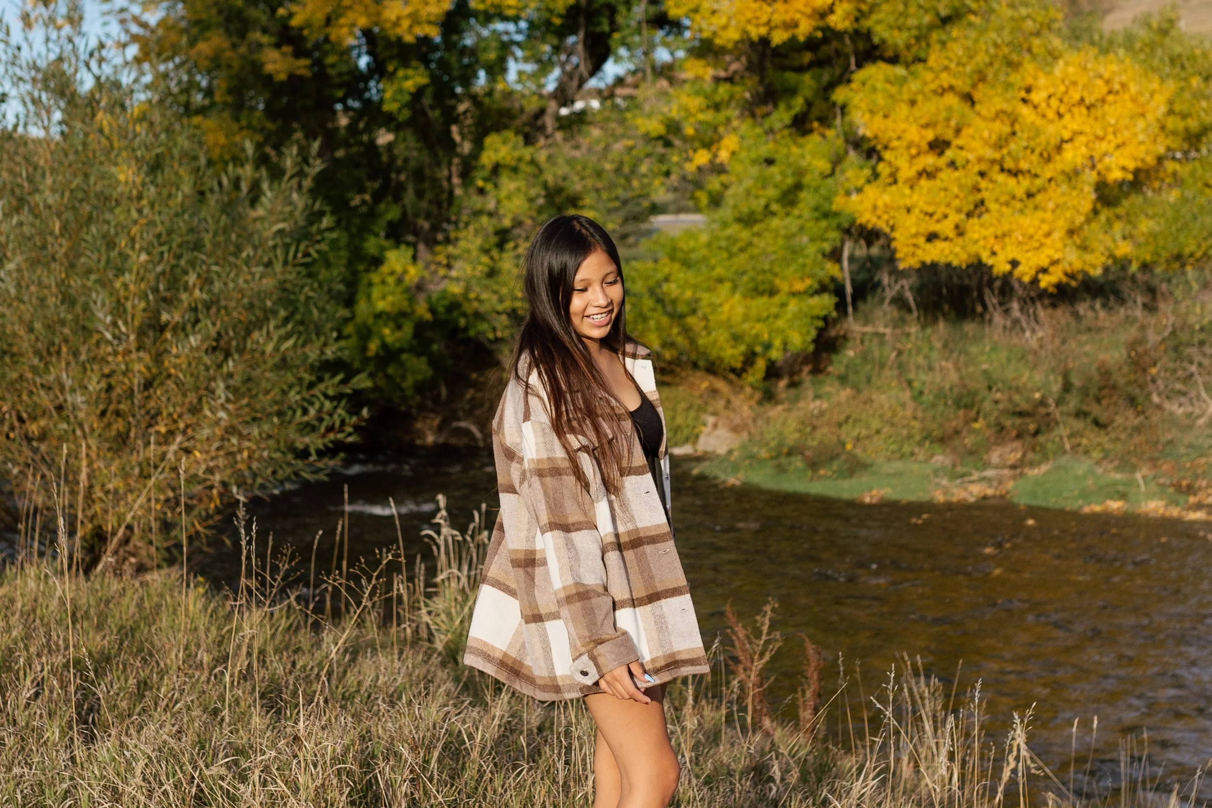 Young woman standing by a river in autumn with yellow and green trees in the background, wearing a plaid jacket and smiling.