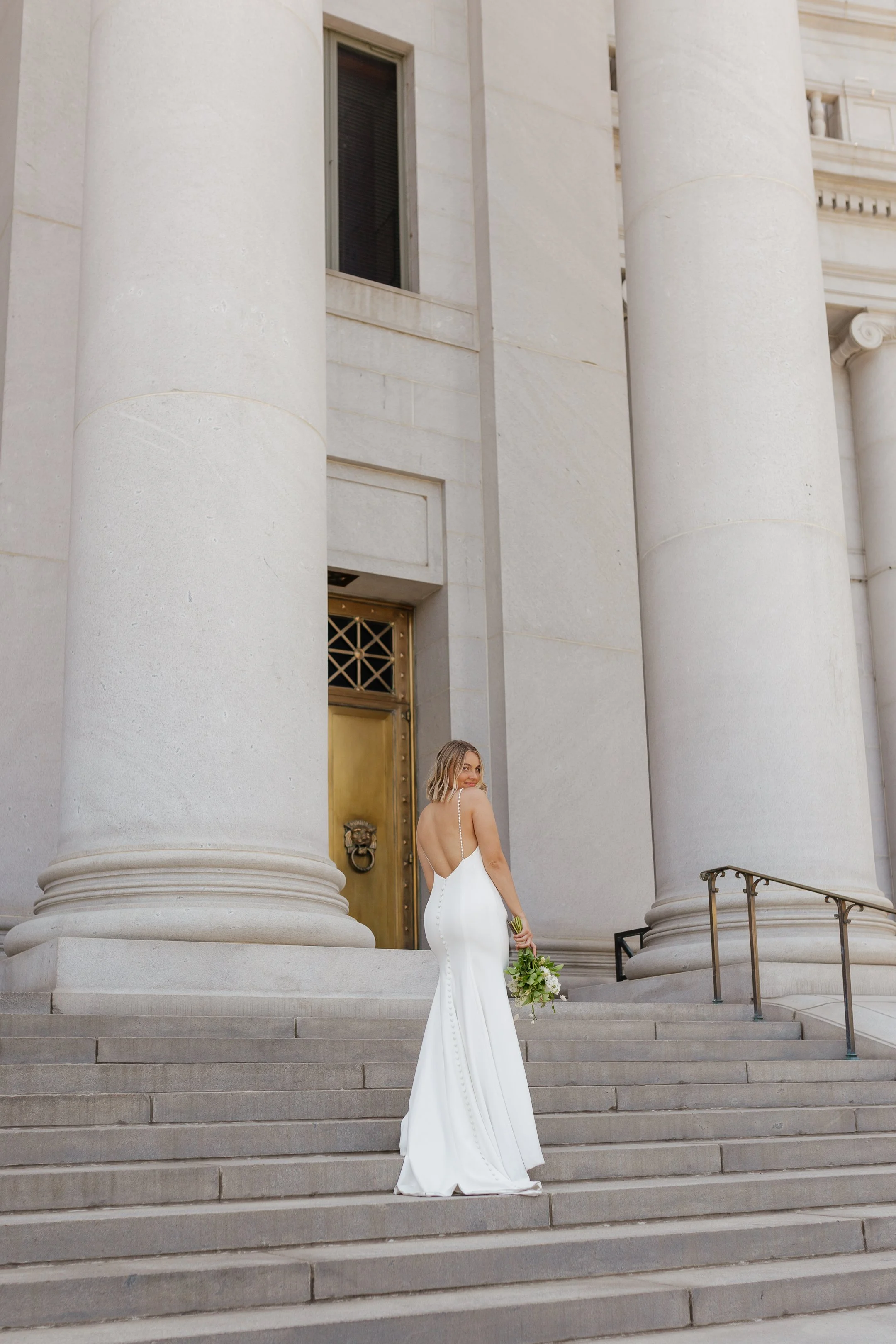 A woman in a white wedding dress holding a bouquet standing on steps in front of a large stone building with tall columns.