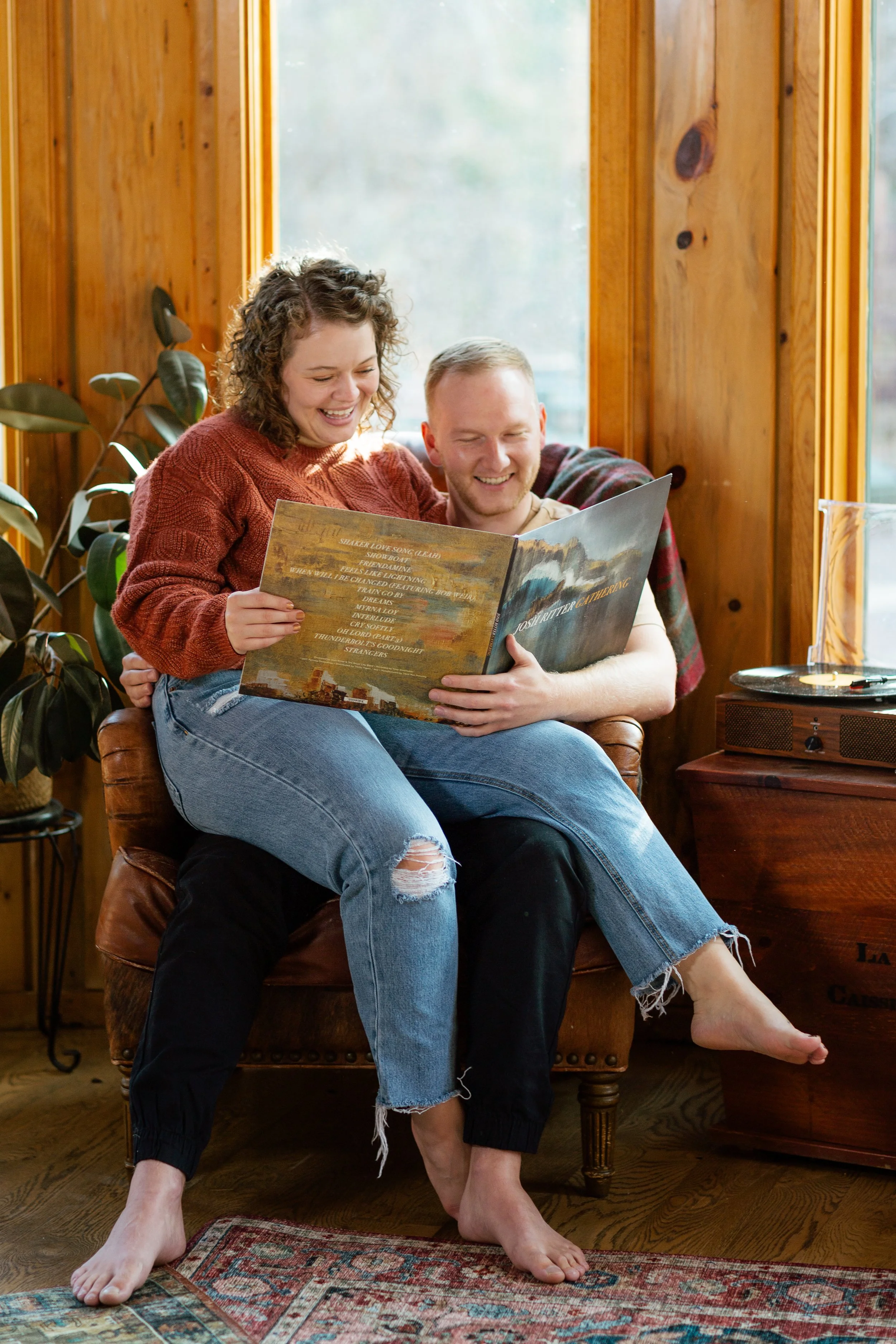 A young woman sitting on a man's lap, both smiling and looking at a music album. The scene is inside a cozy, wooden room with large windows, a carpet, and a potted plant nearby.