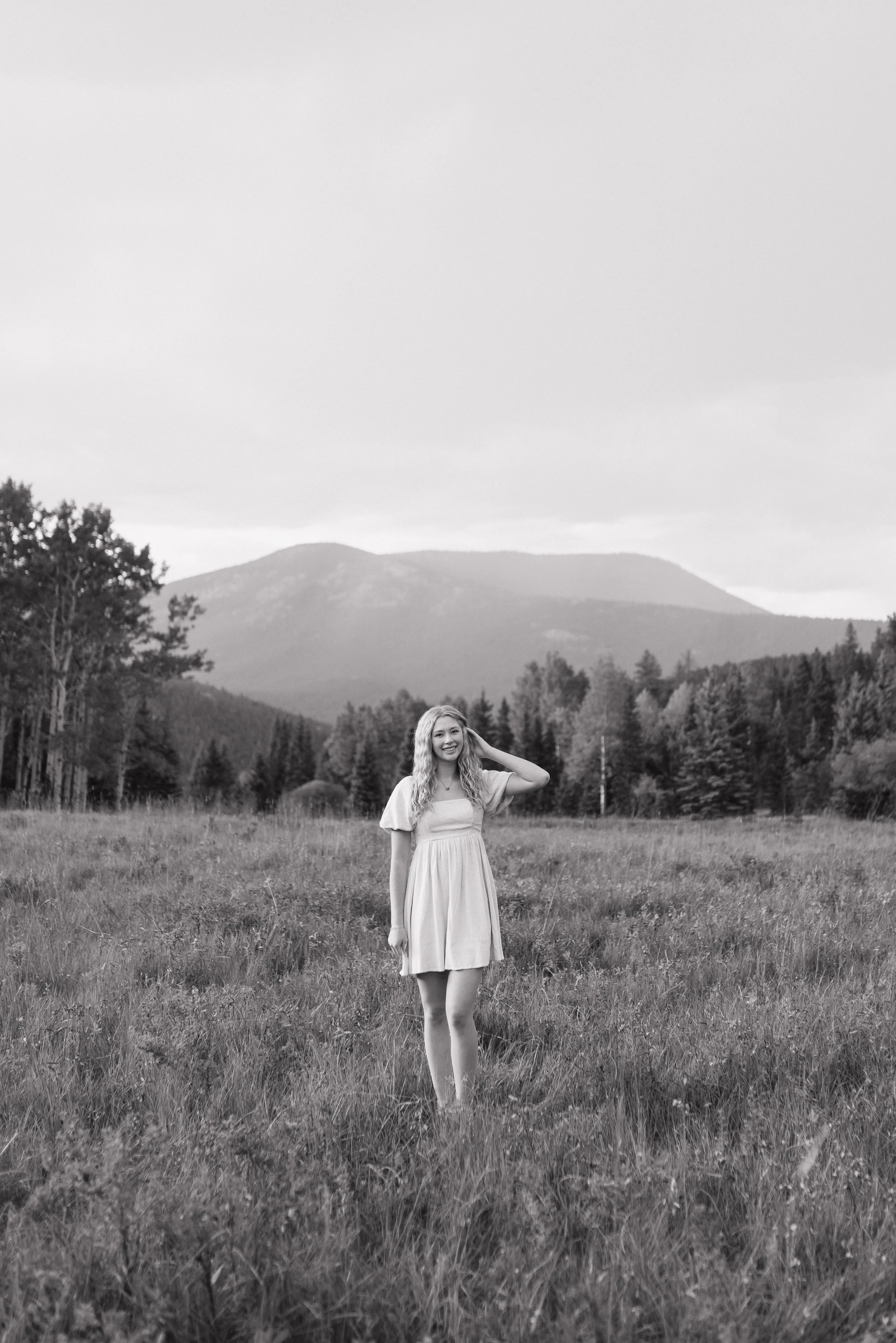 Young woman standing in grassy field with mountains and trees in the background, smiling and touching her hair