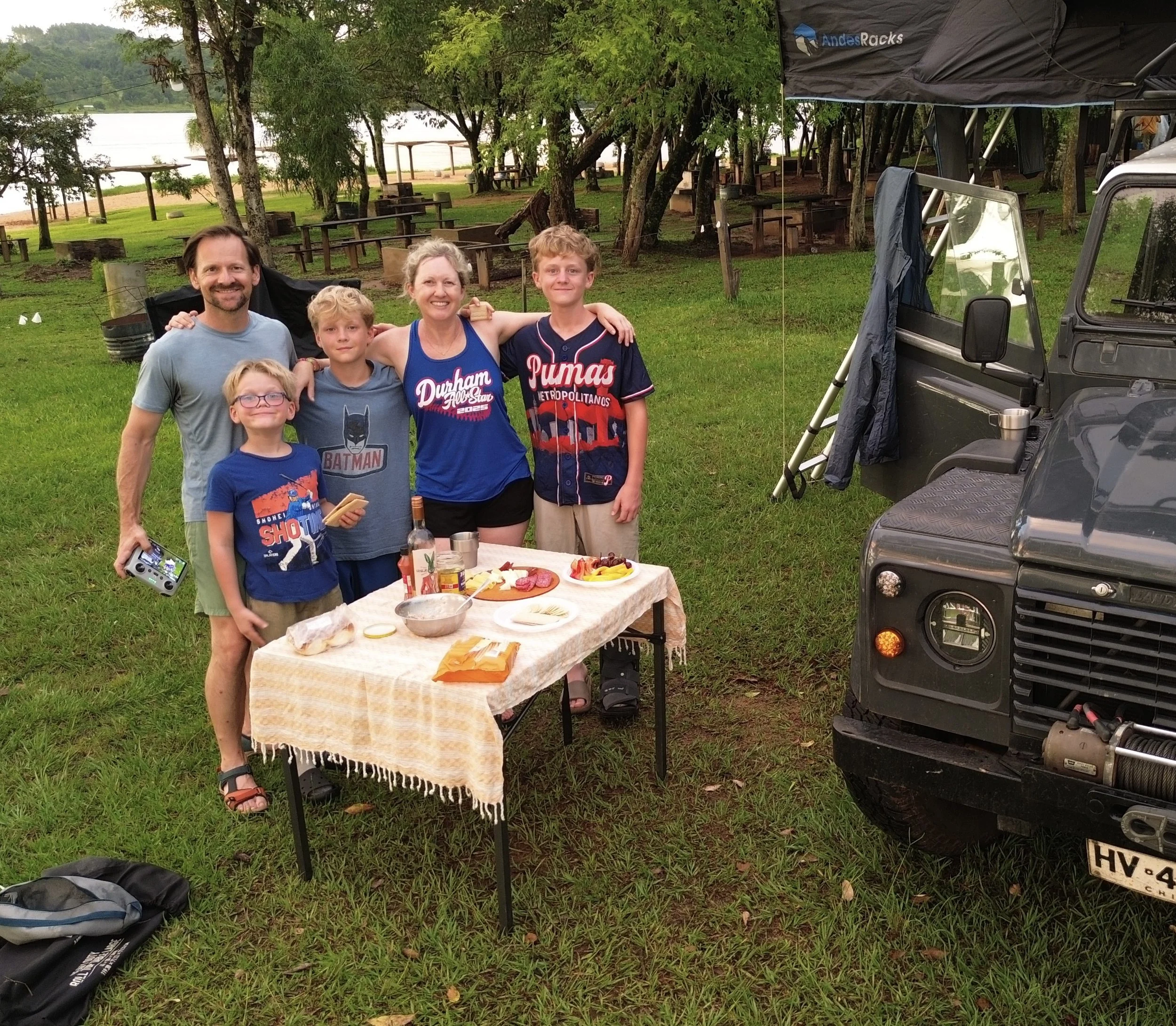 A family of five posing outdoors near a table with food, in a grassy area with trees and a lake in the background, next to a black off-road vehicle and a canopy.