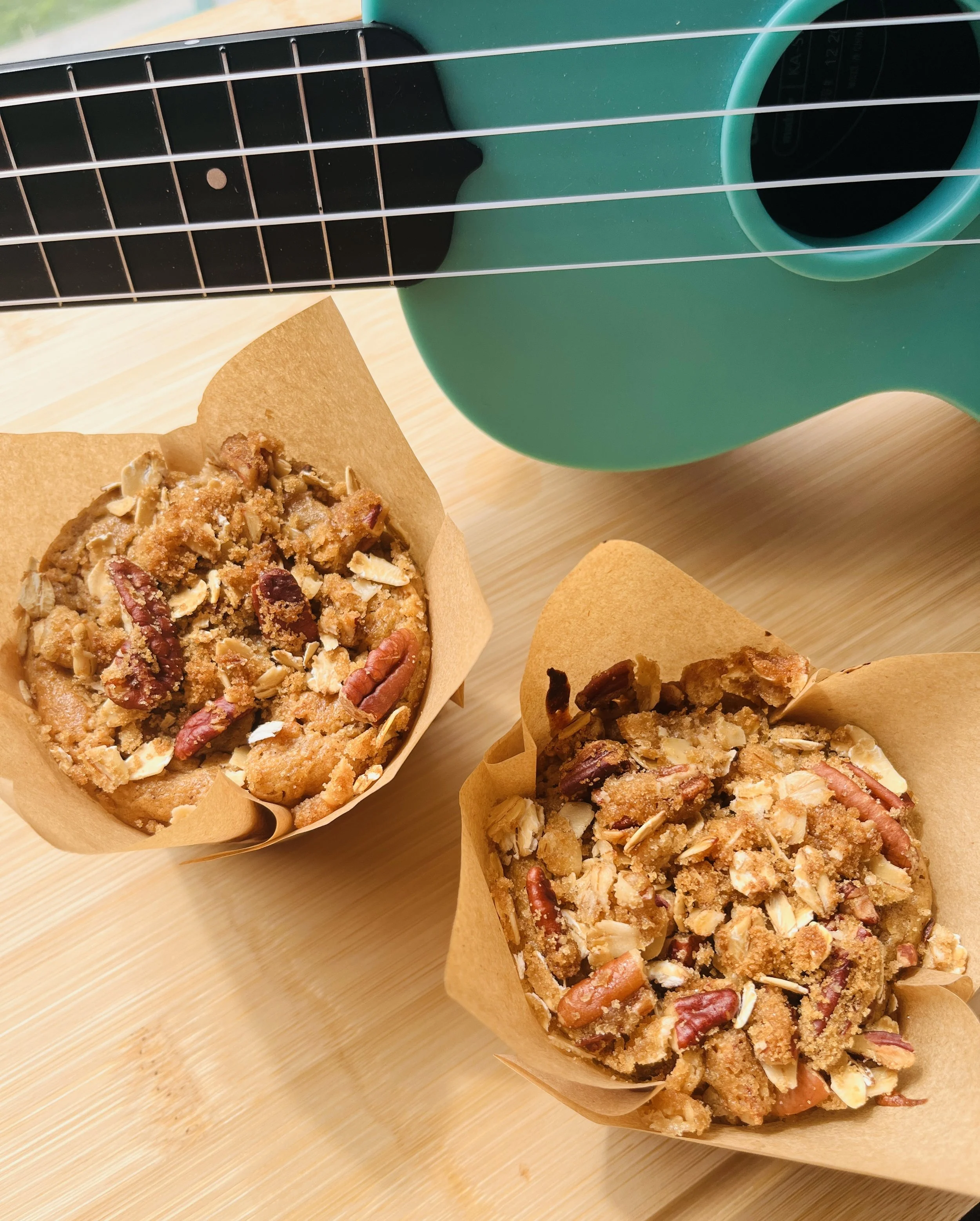 Two whole grain blueberry oat muffins topped with oats and pecans, set on a wooden surface, with a ukulele partially visible in the background.