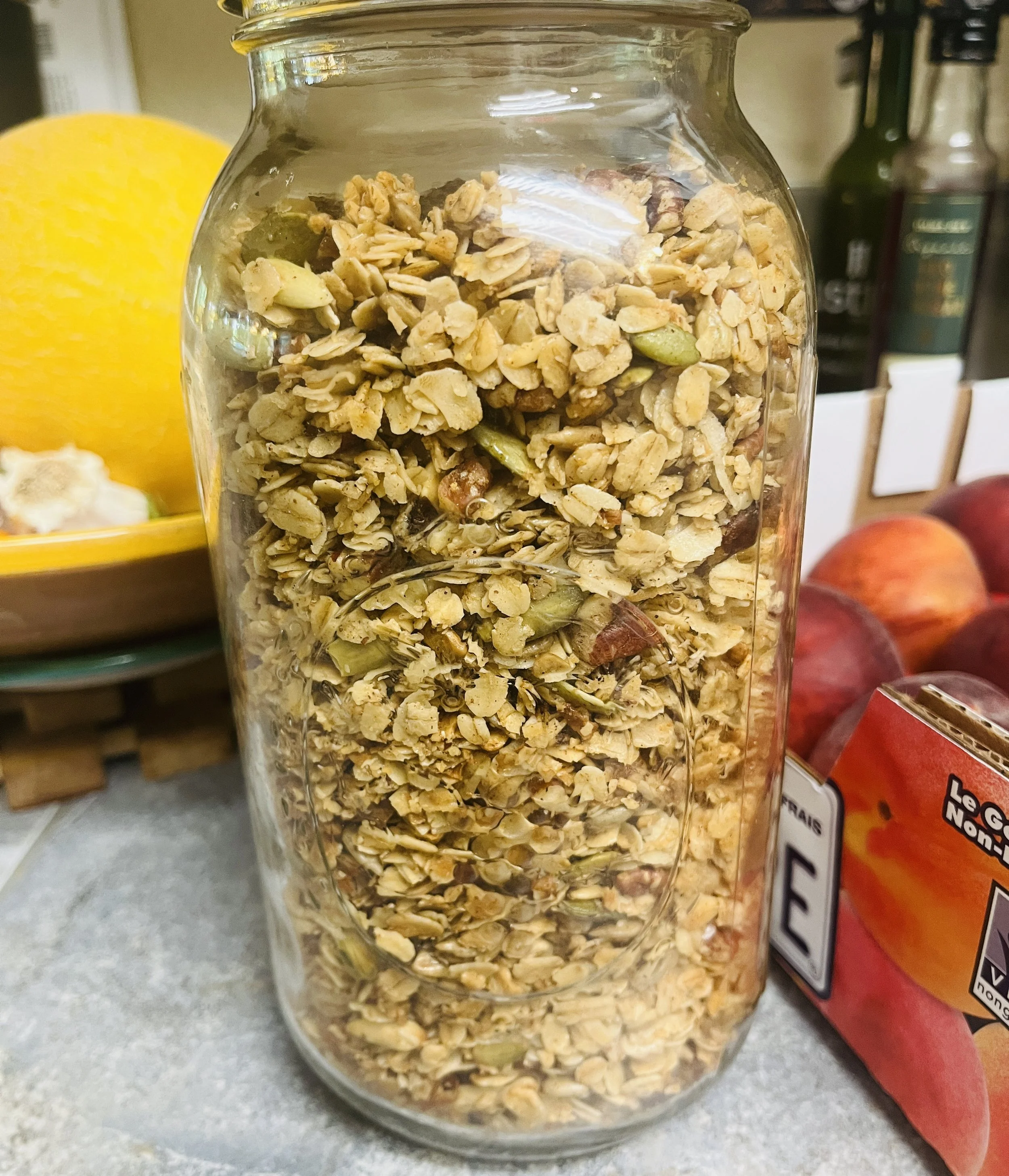 A clear glass jar filled with granola on a countertop, with apples and a yellow fruit in the background.