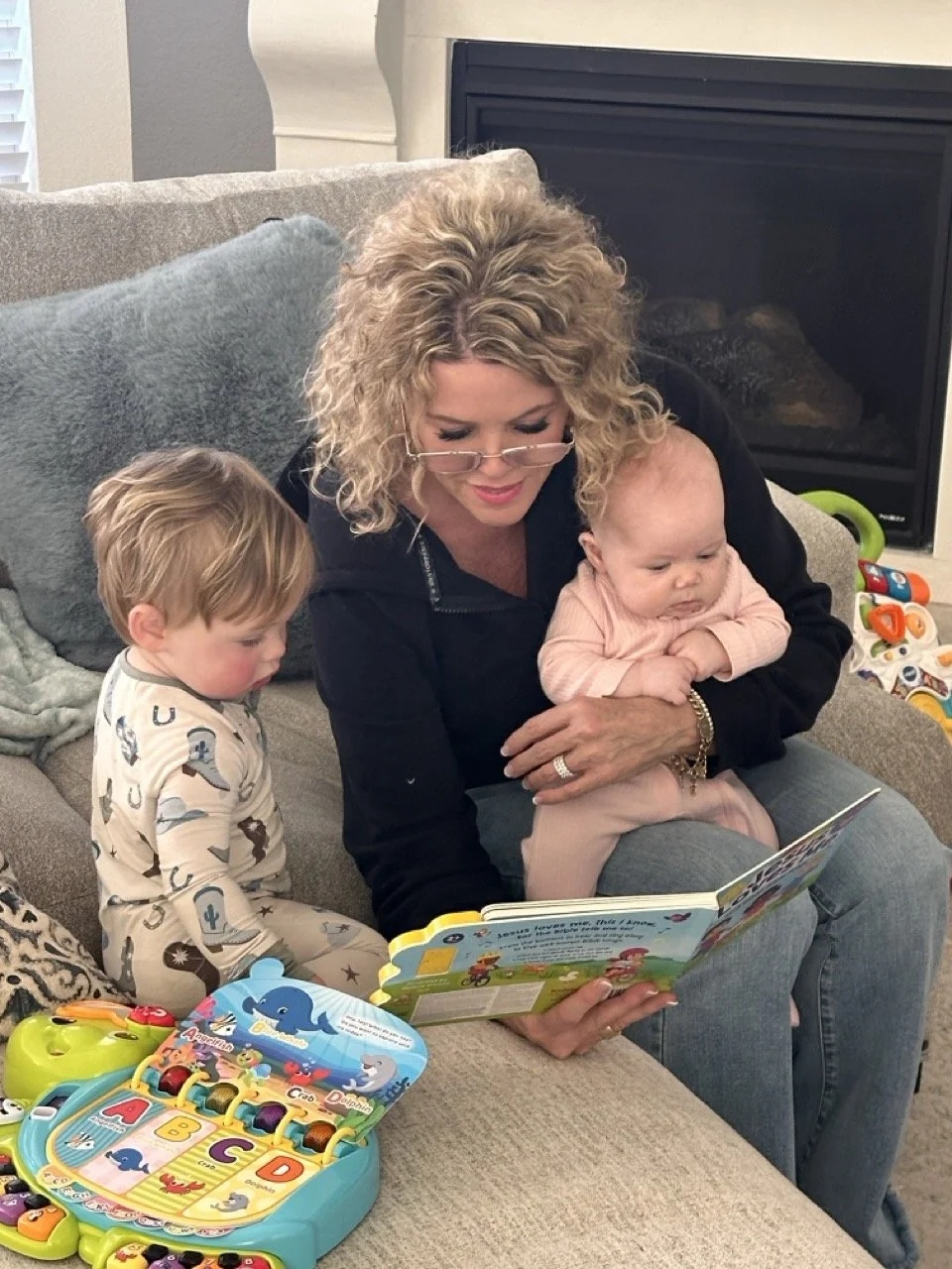 Josie Breaux reads a children's book to her two young grandchildren, sitting on a couch in a living room with toys nearby.
