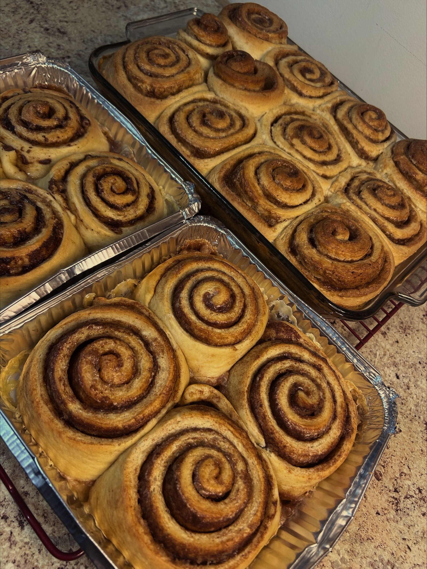 Two trays of freshly baked cinnamon rolls with swirled cinnamon filling, some placed in aluminum foil containers and others in a glass baking dish.
