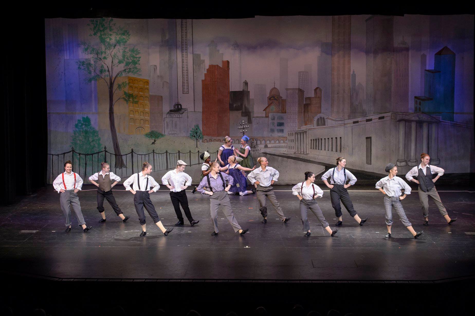 The image shows a group of young dancers performing on stage with a cityscape backdrop, dressed in vintage-style clothing.