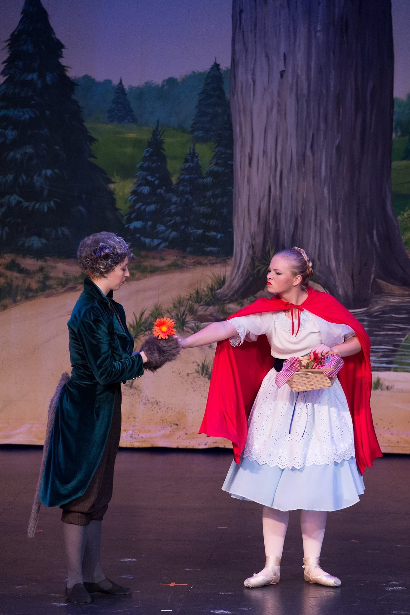 Two women dressed as characters from 'Little Red Riding Hood' on stage, with a painted forest background. One woman is dressed as the wolf, offering her paw with a flower, and the other as Little Red Riding Hood, holding a basket.