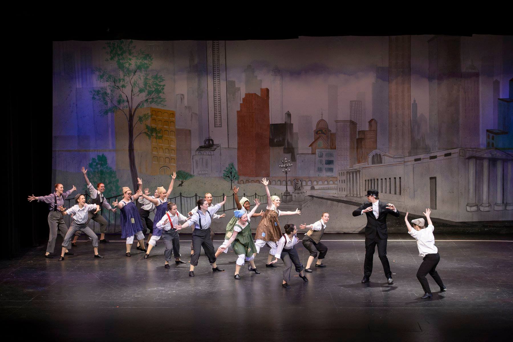 Children performing a dance on stage dressed in period costumes with a cityscape backdrop.
