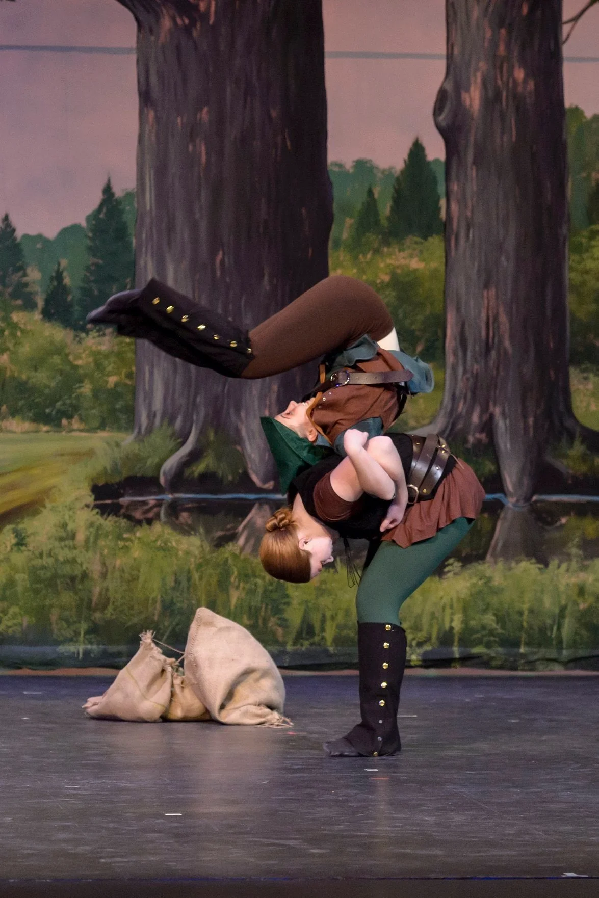 Two performers dressed as fantasy characters engaging in a staged acrobatic act on a stage with a forest-themed backdrop, with burlap sacks on the floor near them.