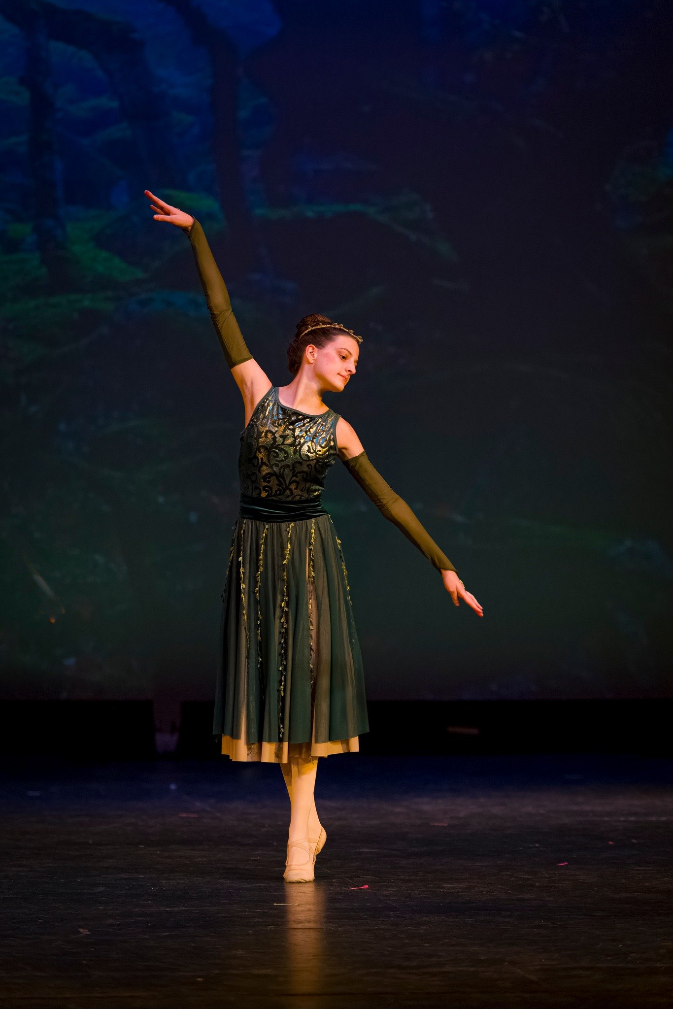 A ballerina performs on stage, wearing a dark green and gold costume with long gloves and ballet shoes, against a dark background.