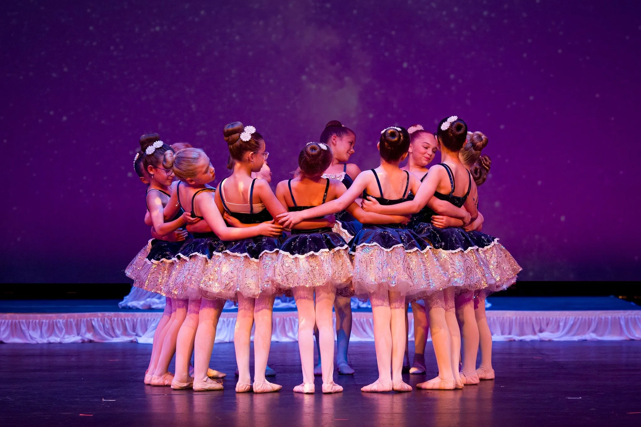 A group of young ballet dancers in tutus and ballet slippers are standing in a circle on stage, embracing each other in a huddle. They are wearing black leotards with sparkly skirts and white floral headbands. The background is purple, and the stage is decorated with a pink ruffled curtain at the front.