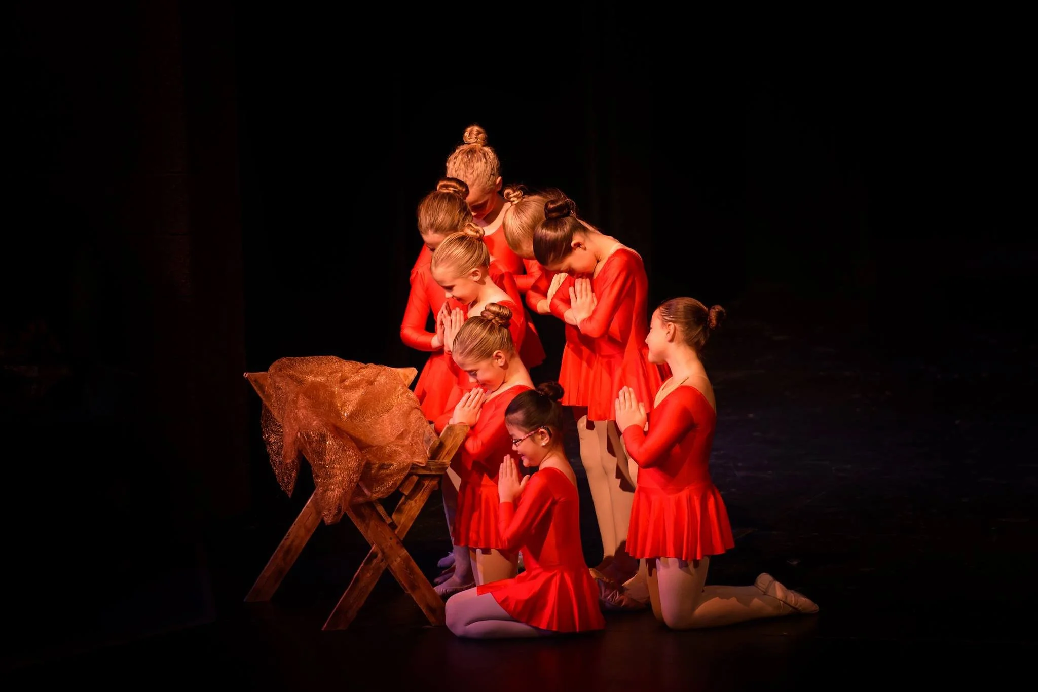 Children dressed in red costumes kneeling and standing with hands in prayer position in front of a manger with a gold star, shaped like a nativity scene on a dark stage.