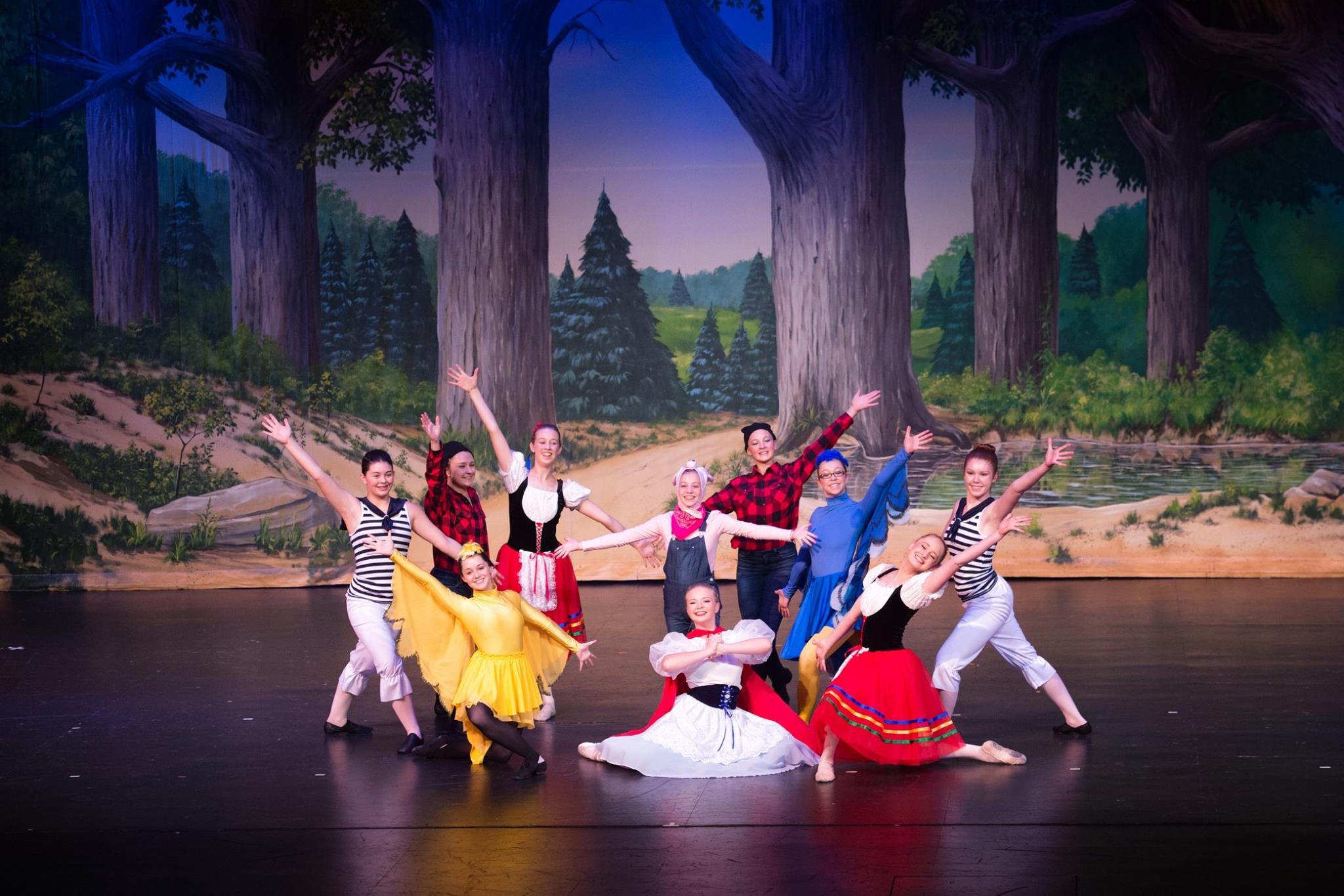 A group of children dressed in colorful costumes performing a dance on stage with a painted forest background.