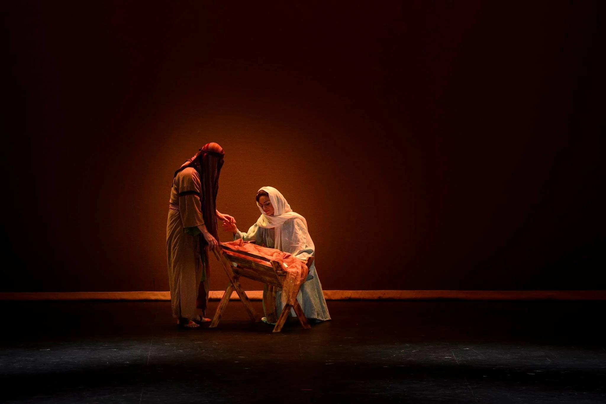 Two women dressed in historical or biblical clothing, one kneeling and the other standing, are gathered around a manger with a cloth, part of a stage performance or nativity scene, with a dark background.