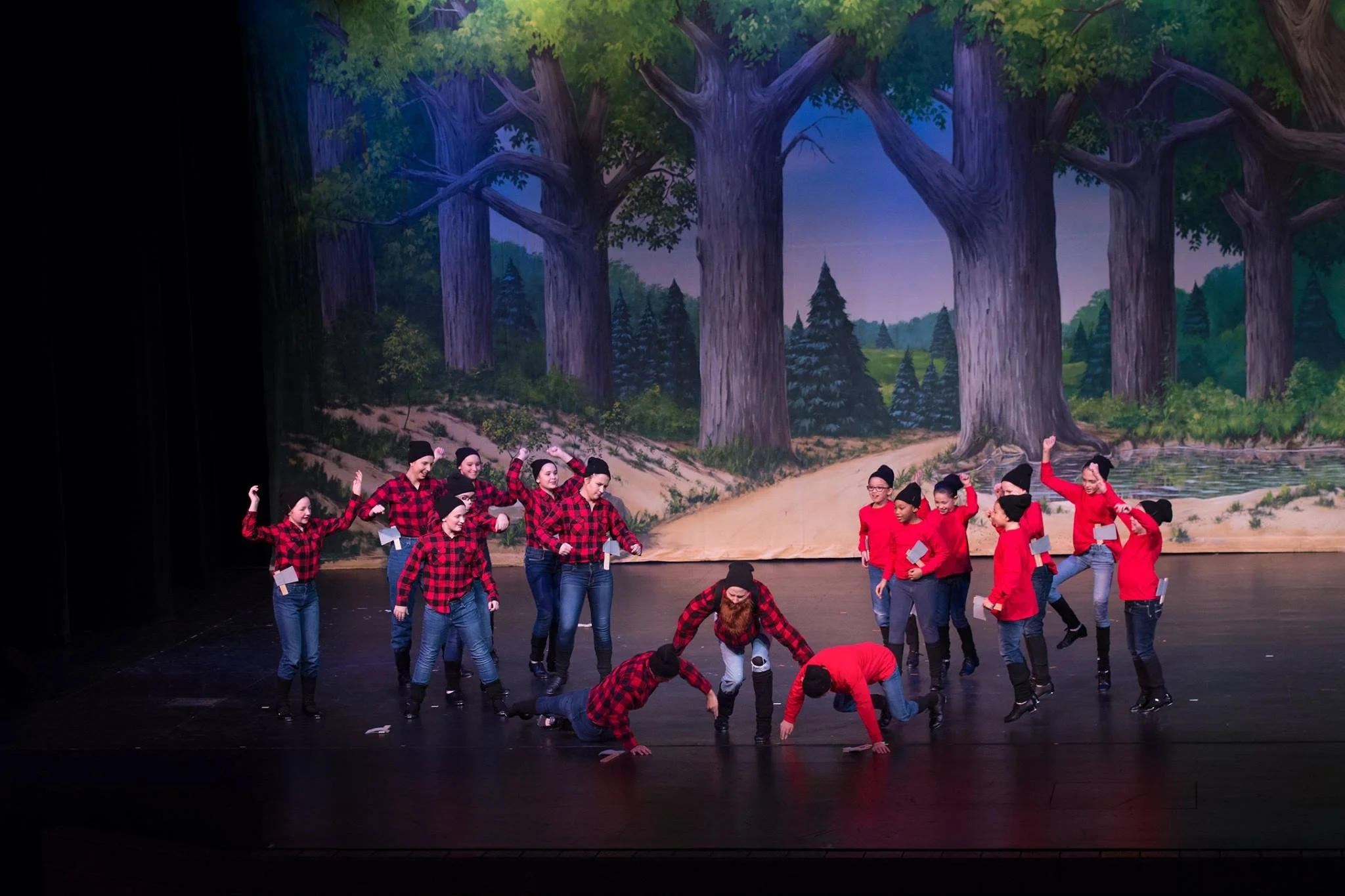 Children performing a stage play or dance in front of a painted forest backdrop, dressed in plaid shirts, jeans, and black hats.