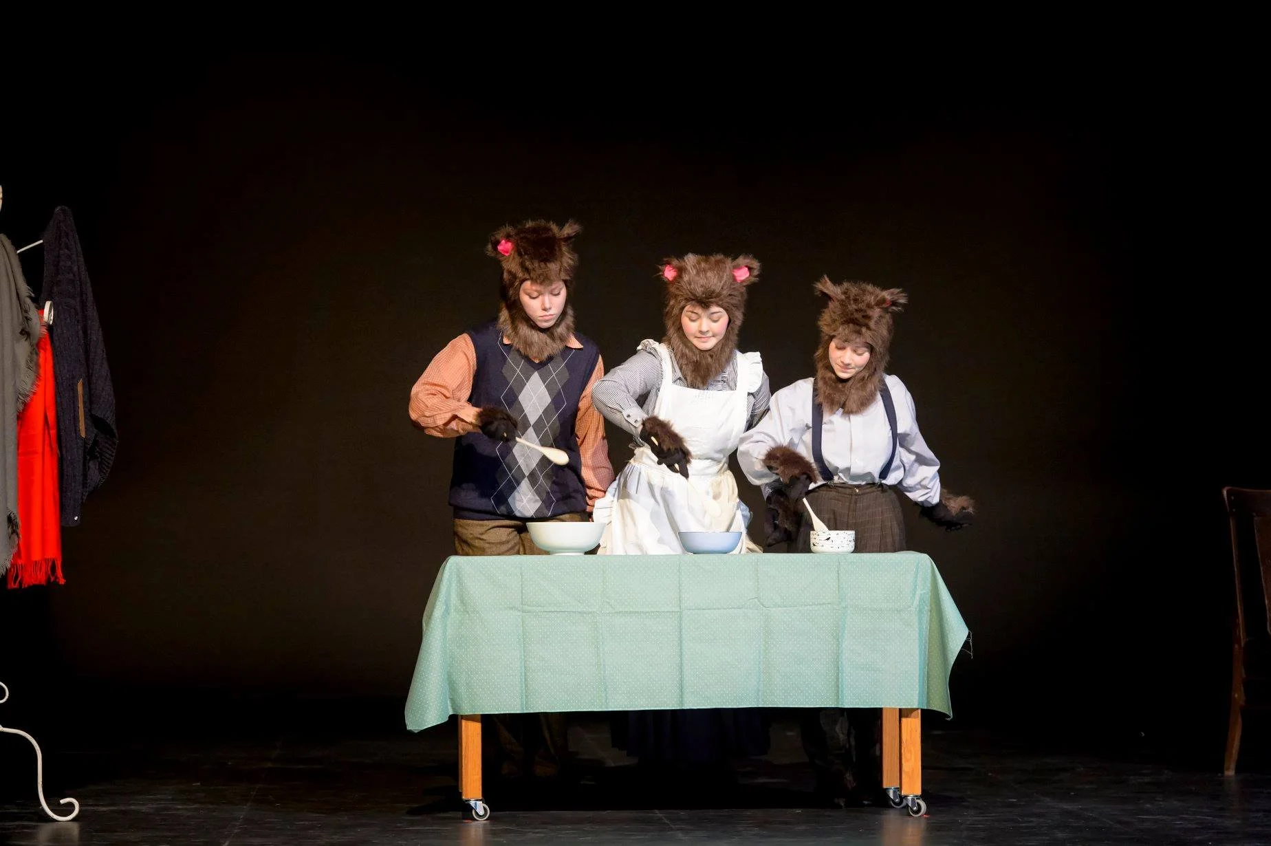 Three children dressed as wolves stand behind a table on stage, stirring bowls as part of a theatrical performance with a black background.