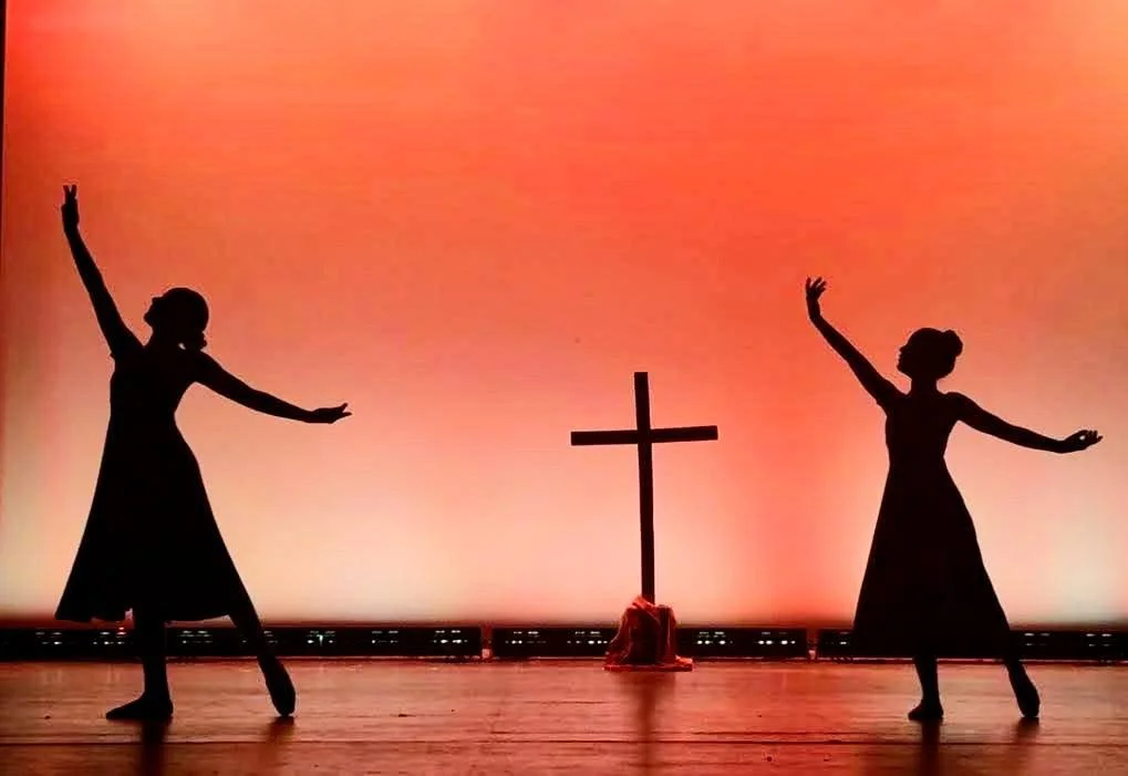 Two women in dresses performing a dance on stage with a cross and cloth in the background.