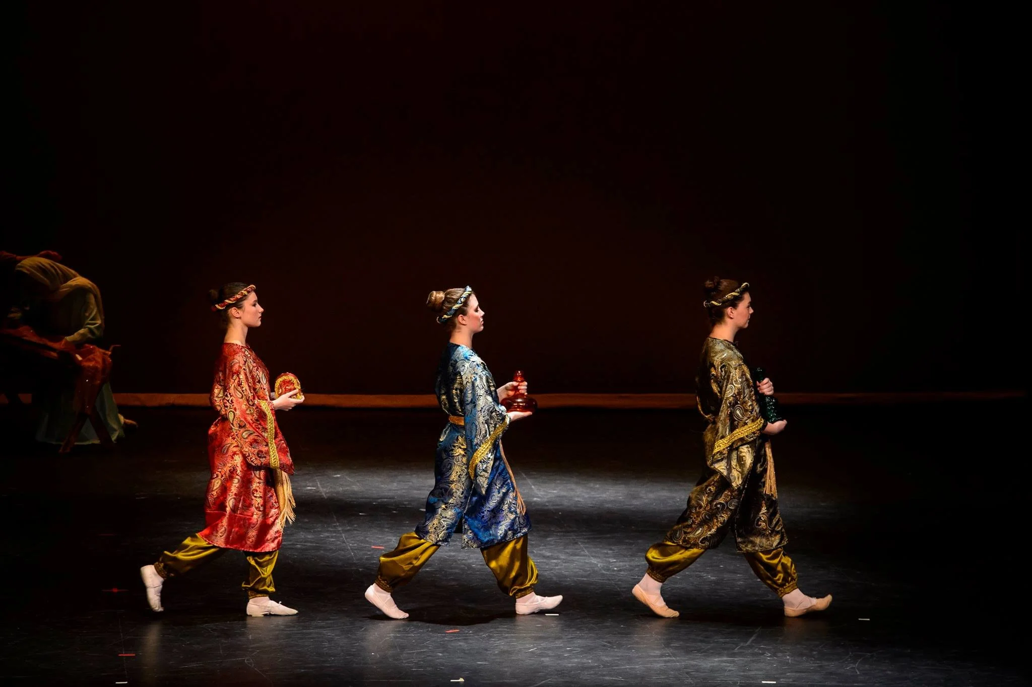 A group of ballet dancers dressed in colorful traditional costumes walking in a line on stage during a performance.