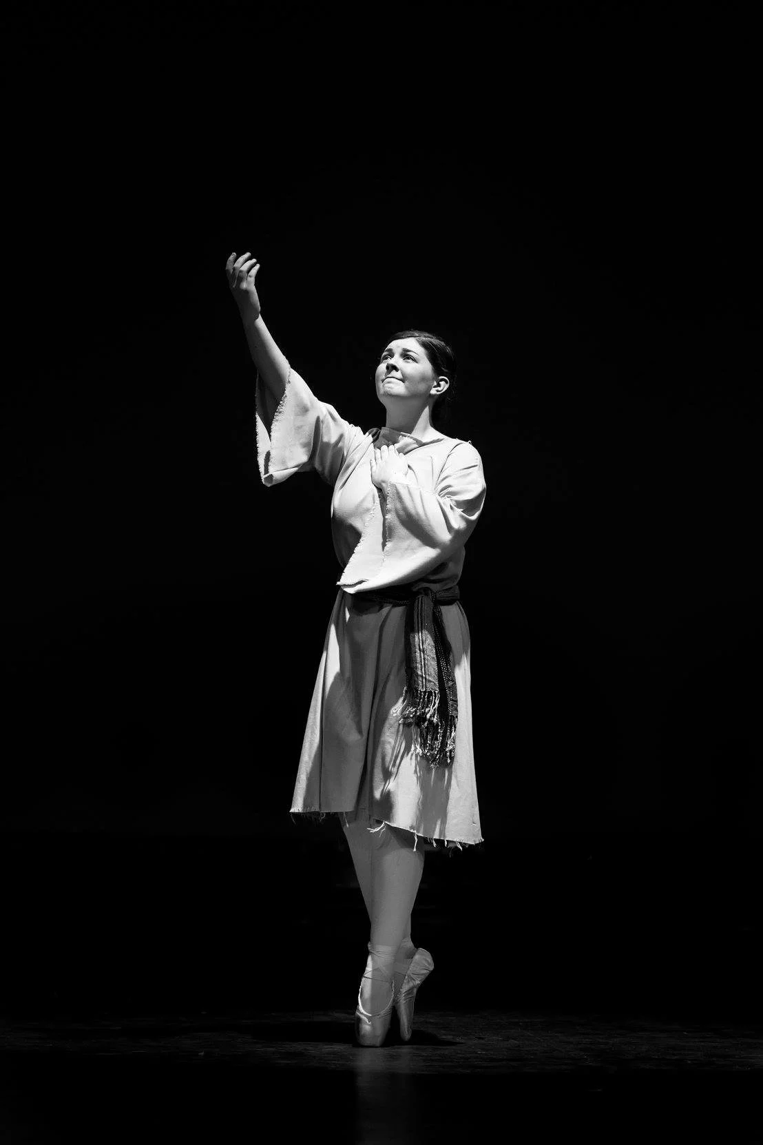 A female ballet dancer performing on stage in a costume, her right arm extended upward, and she is on pointe in ballet slippers, captured in a black and white photograph.