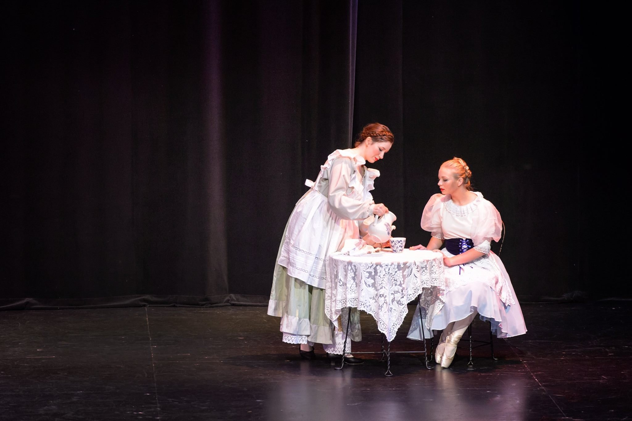 Two women dressed in vintage costumes standing at a table with a lace tablecloth, one pouring tea into a cup, set against a dark stage background.