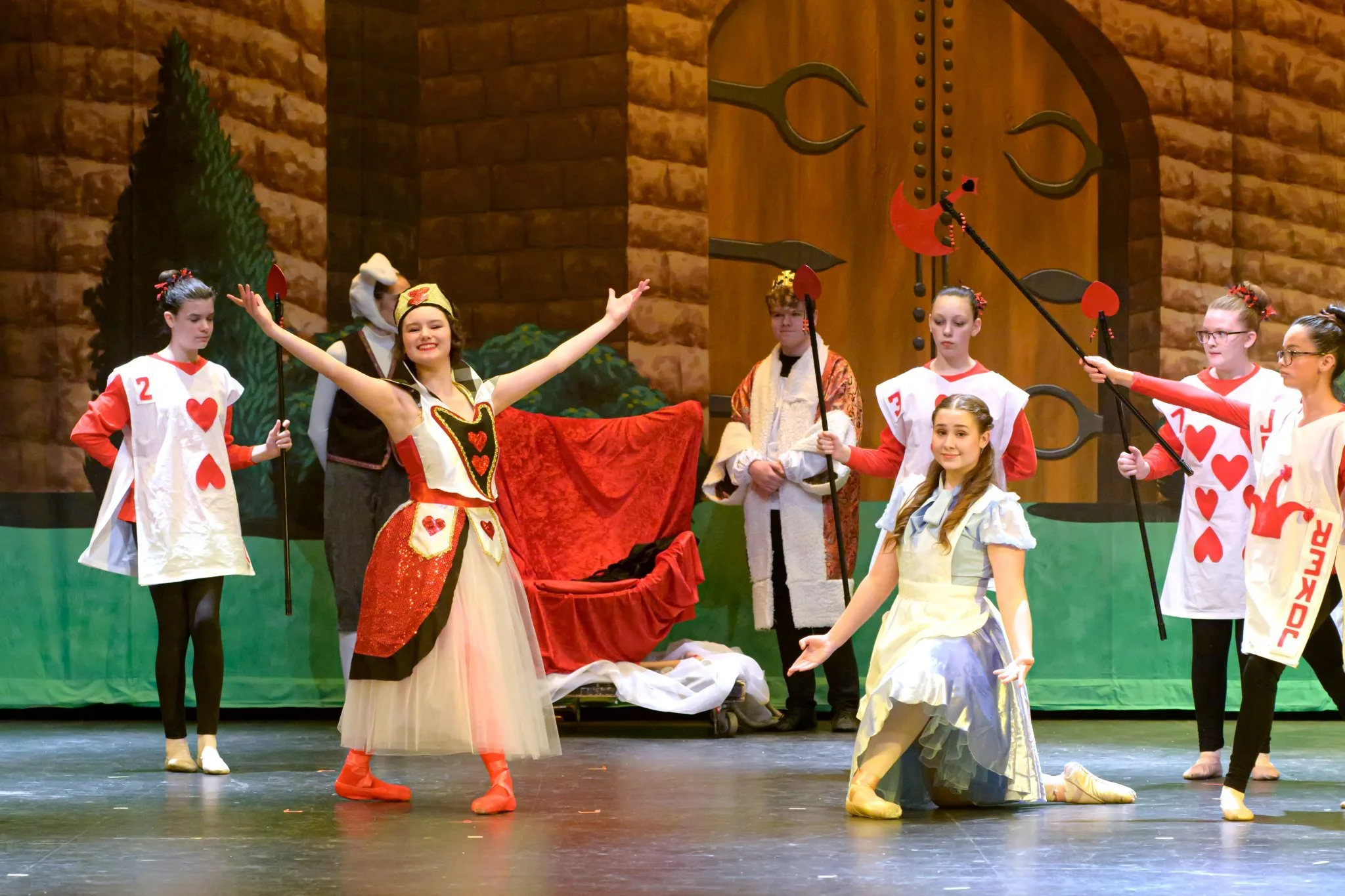 Children in playing costumes performing on stage in a theatrical play. Some are dressed as playing cards, and the background shows a set resembling a castle with trees and a wooden door.