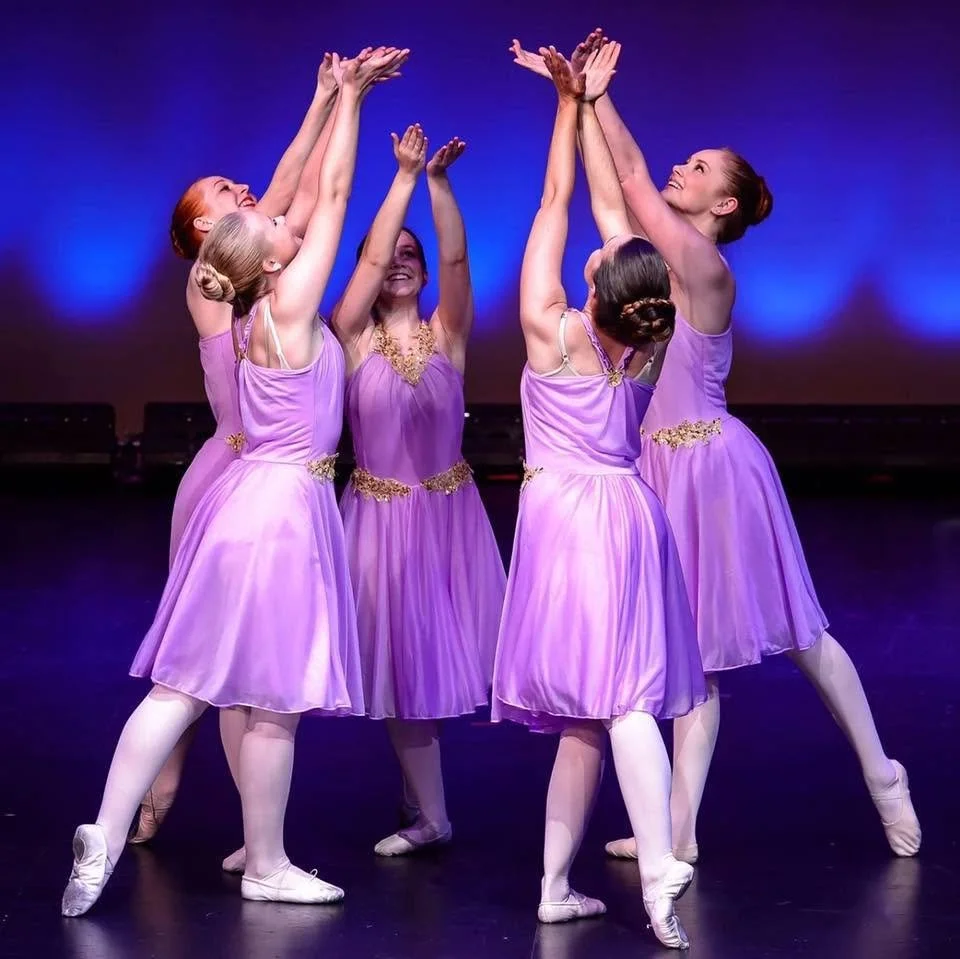 Group of five female ballet dancers in purple costumes performing on stage with blue lighting.