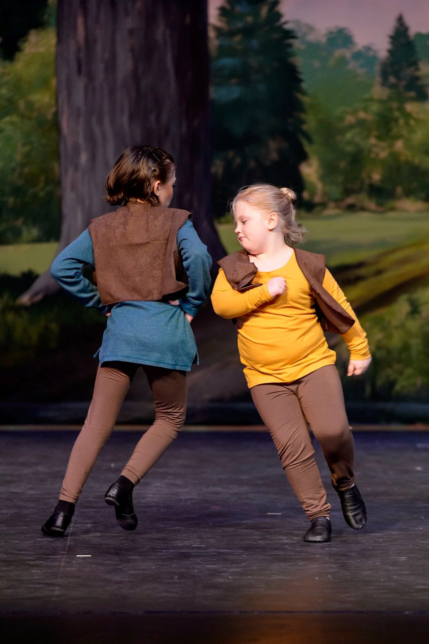 Two young women dressed as dancers in brown and yellow costumes, performing on a stage with a backdrop of trees and a large tree trunk.