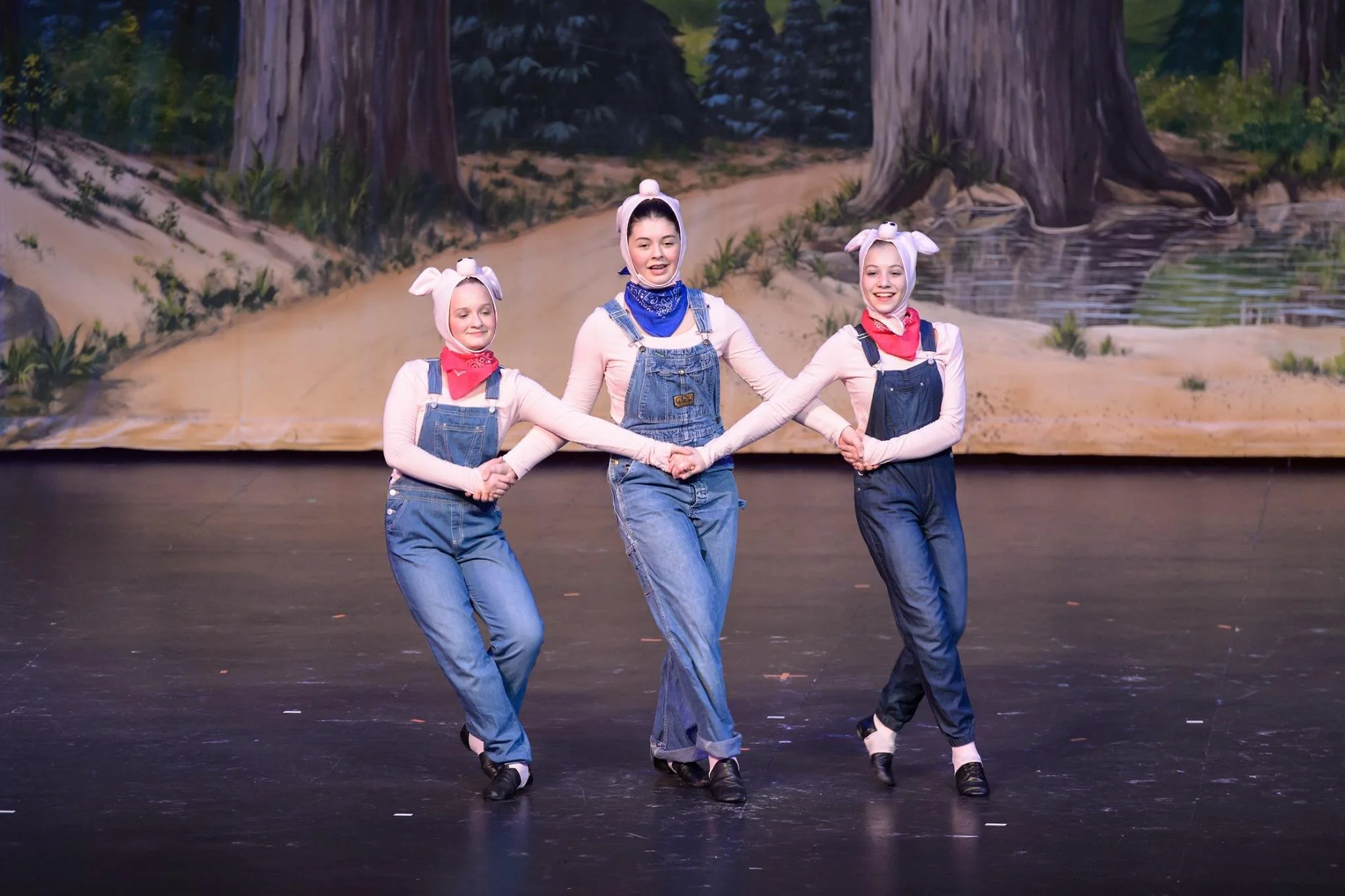 Three young women in costume with pink bunny ears headbands and bandanas, holding hands and dancing on stage with a forest backdrop.