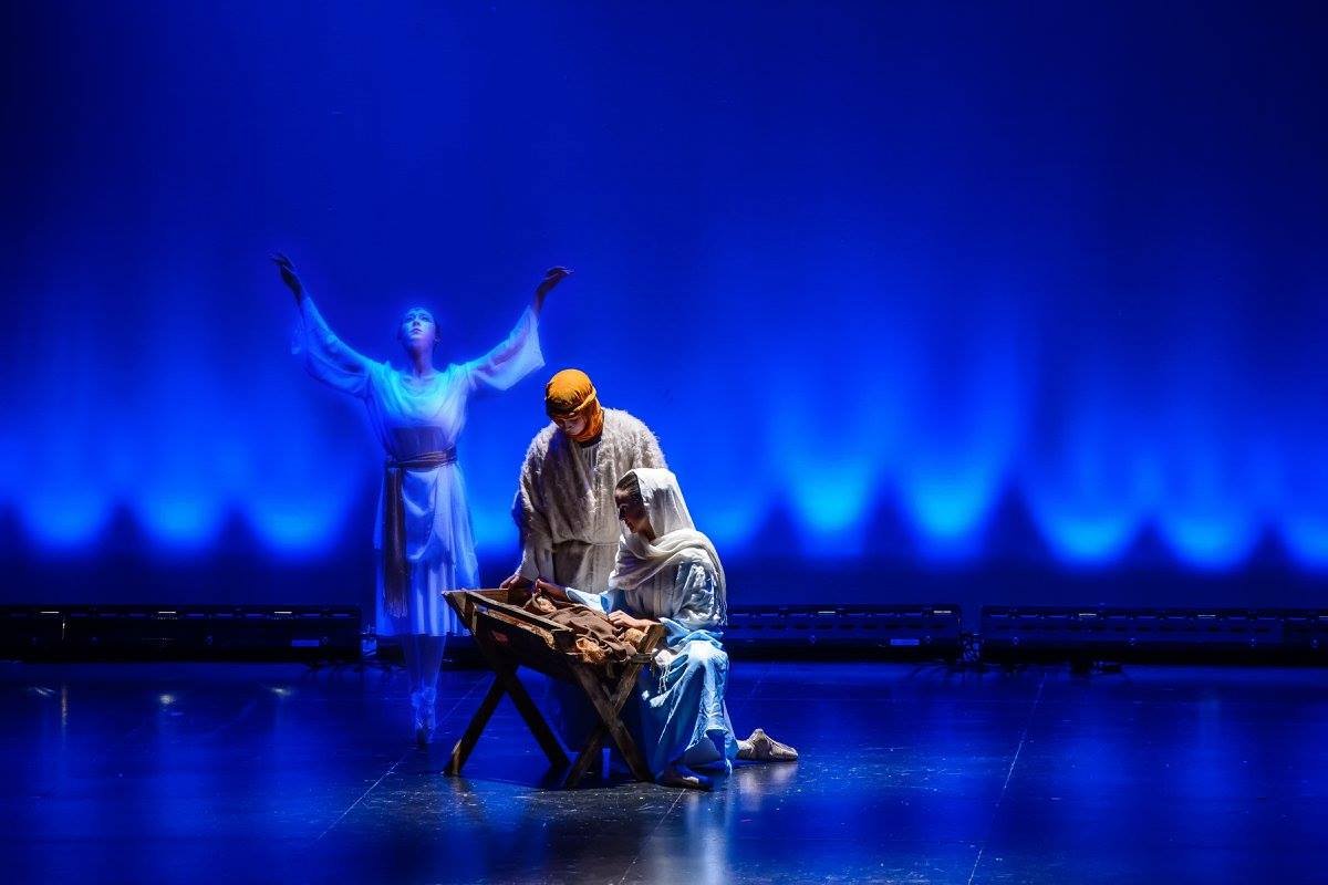 Three performers on stage in a nativity scene, with one figure in blue robes with arms raised, and two others dressed in traditional biblical attire; one playing a harp-like instrument.