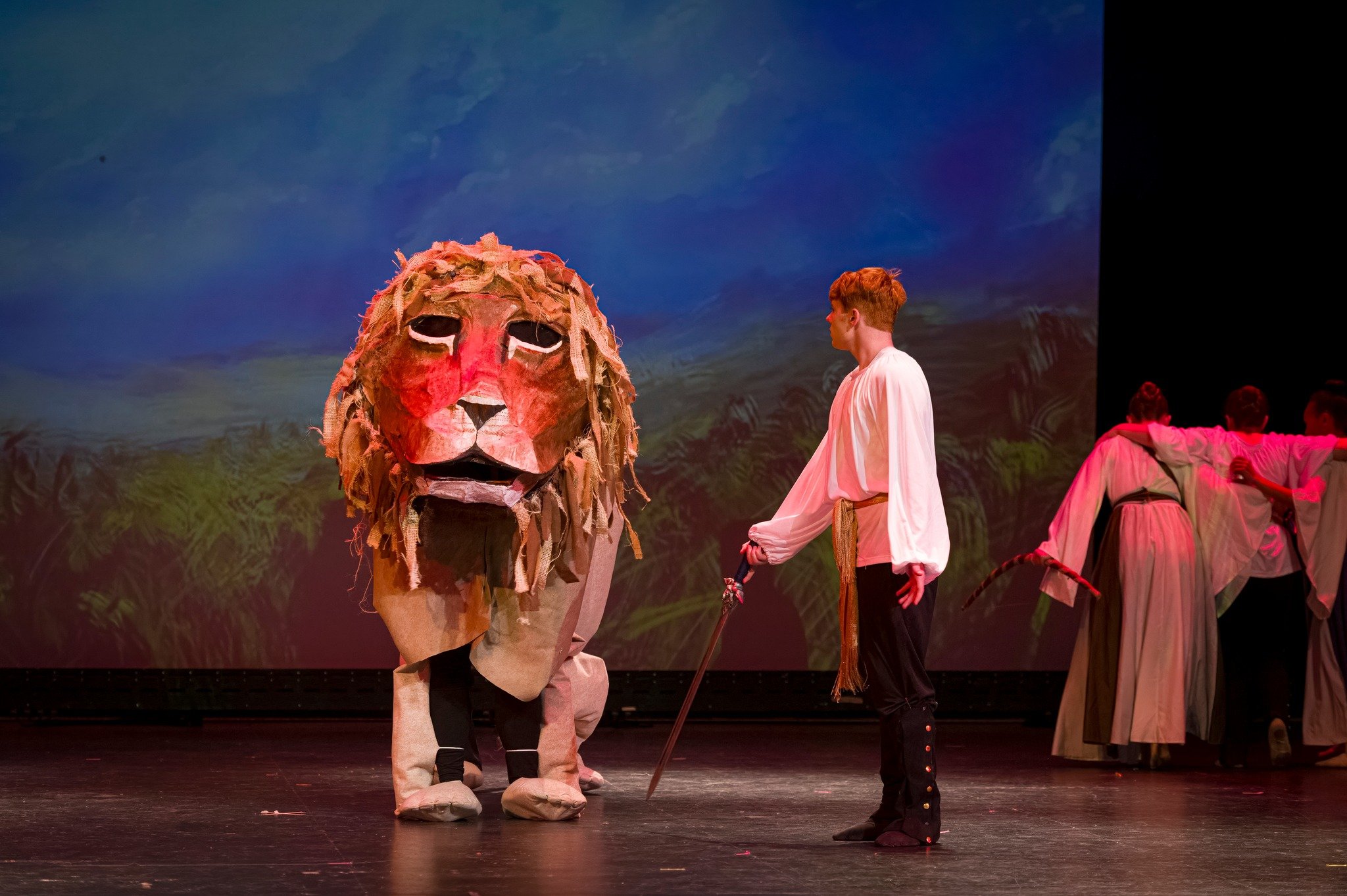 Stage performance with performers in costumes. One performer wears a large lion costume mask with a mane made of fabric strips, another wears a white costume with medieval style trousers, and there are additional performers in the background dressed 