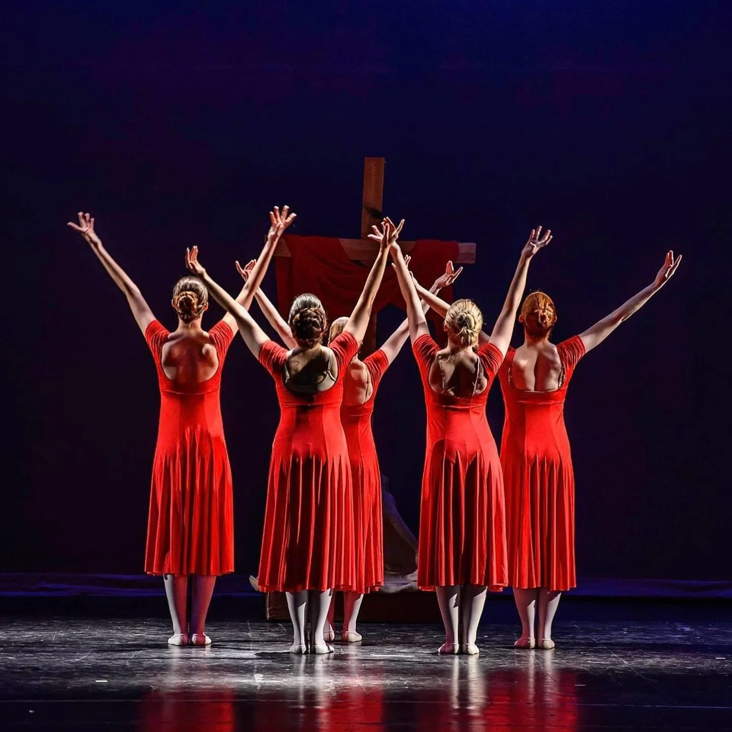 Five ballet dancers dressed in red dresses perform on stage with a large cross in the background.