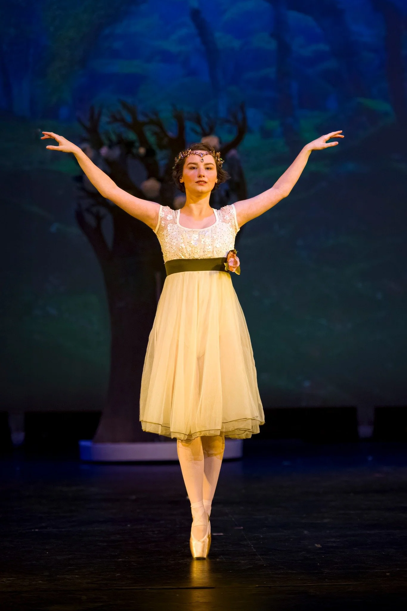 Ballet dancer in a yellow dress performs on stage, wearing pointe shoes and a floral headpiece, with a forest scene projected in the background.