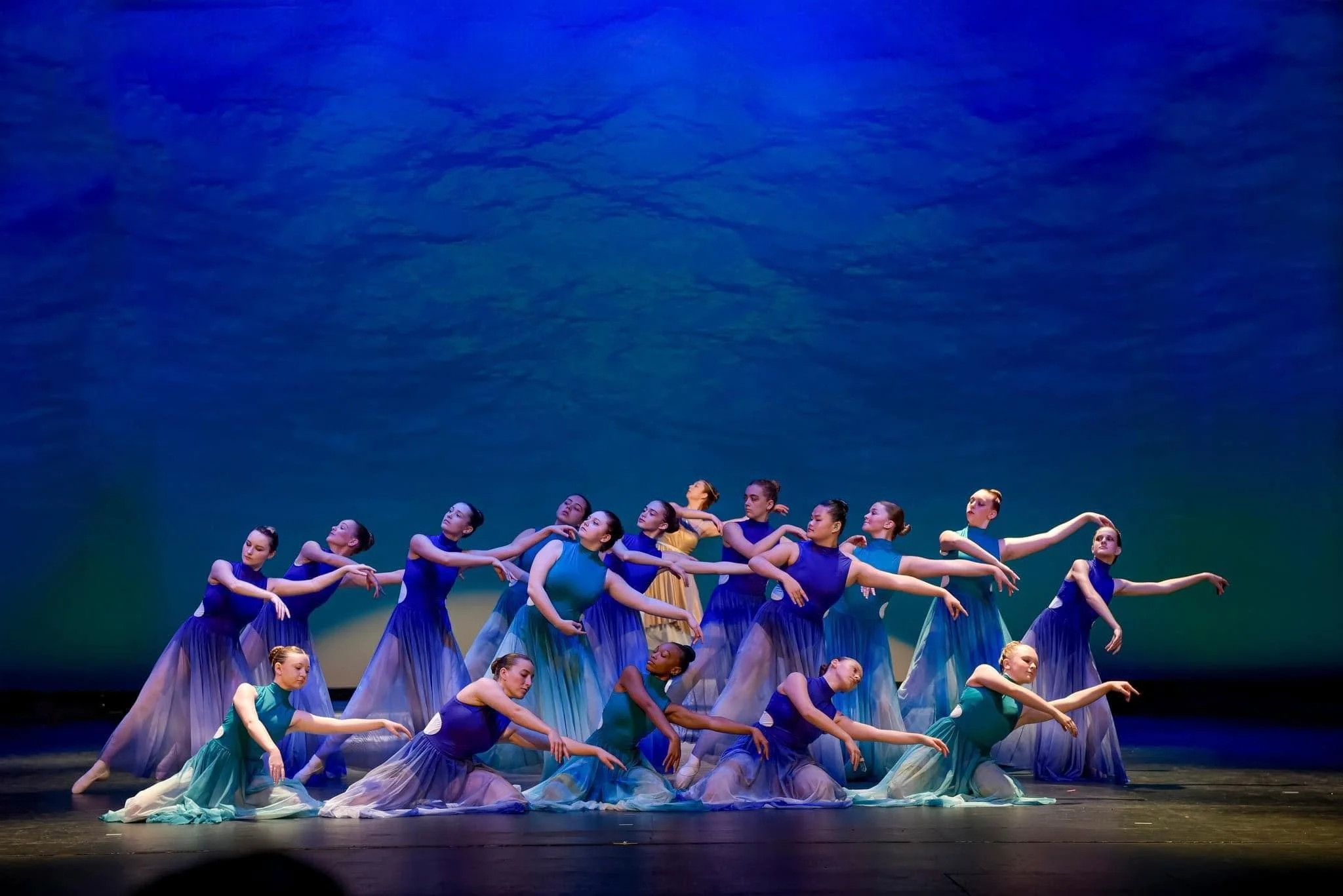 Group of dancers on stage wearing blue, green, yellow, and other colored costumes, performing a ballet or contemporary dance.