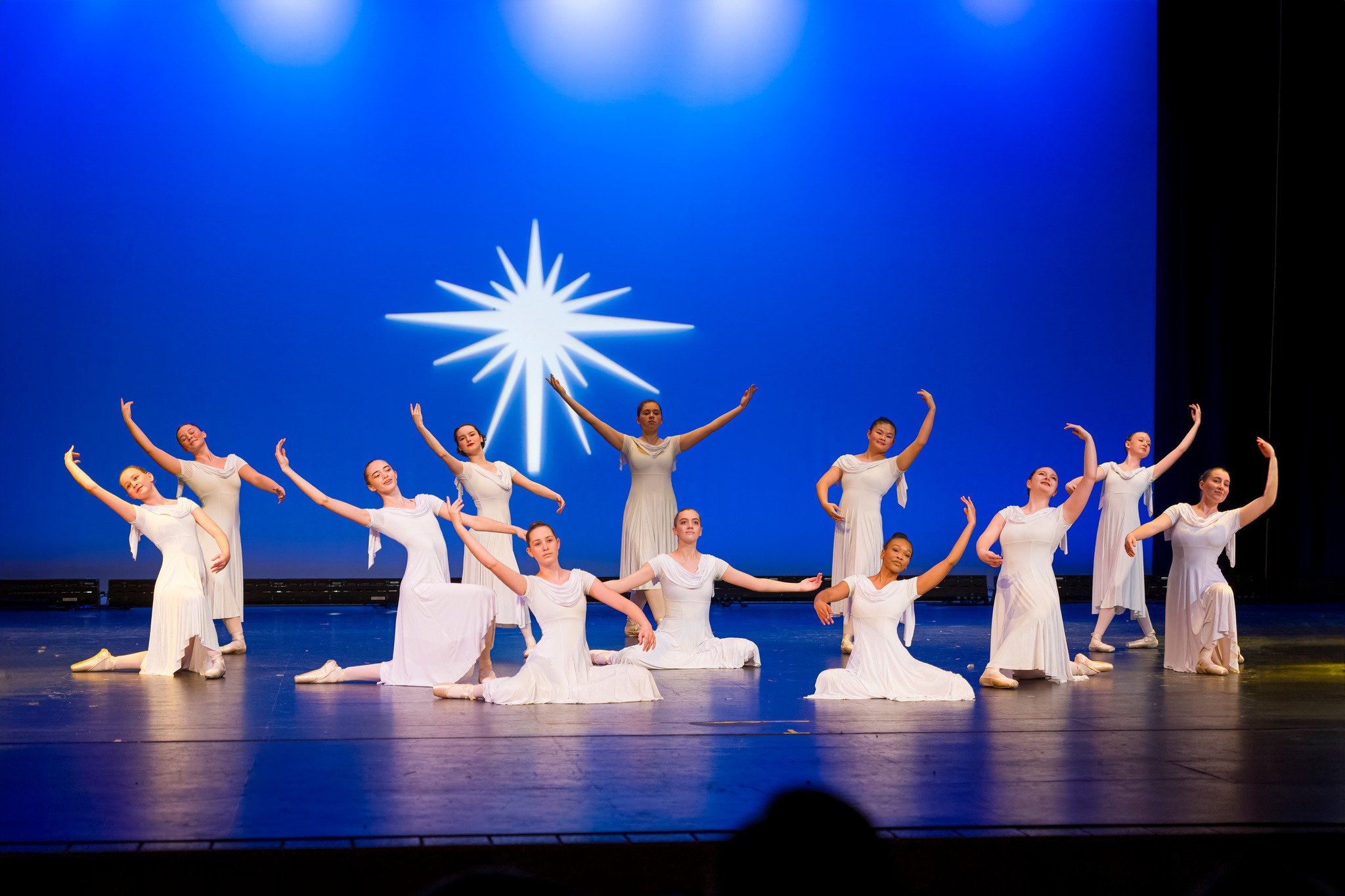 A group of ballet dancers in white costumes performing on stage with a blue background and a white star-shaped light projection.