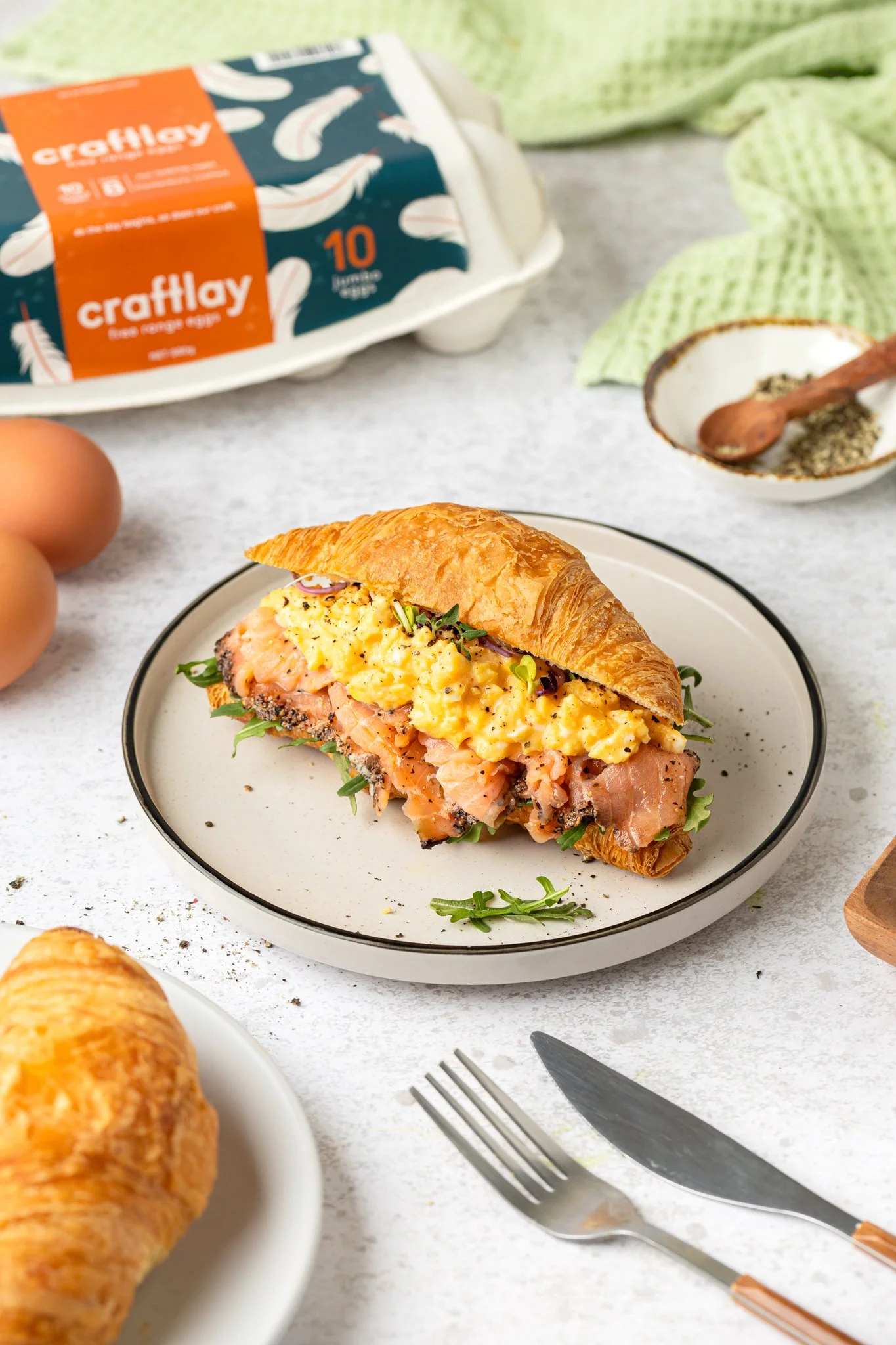 A croissant sandwich with scrambled eggs, smoked salmon, and greens on a white plate, along with eggs, a package of craftily eggs, and a bowl of salt and pepper on a light-colored table.