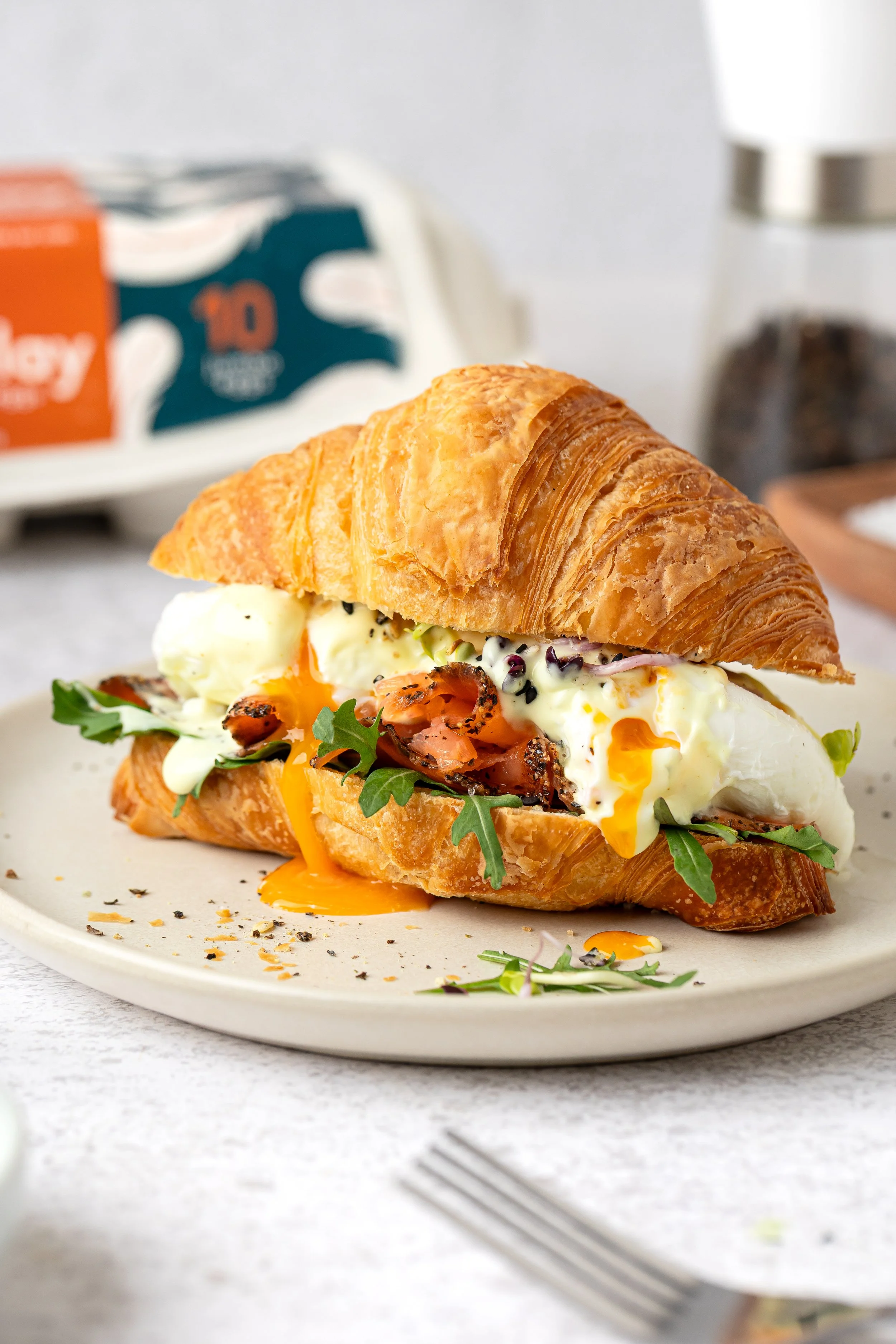 Croissant sandwich with eggs, smoked salmon, arugula, and black pepper on a white plate.